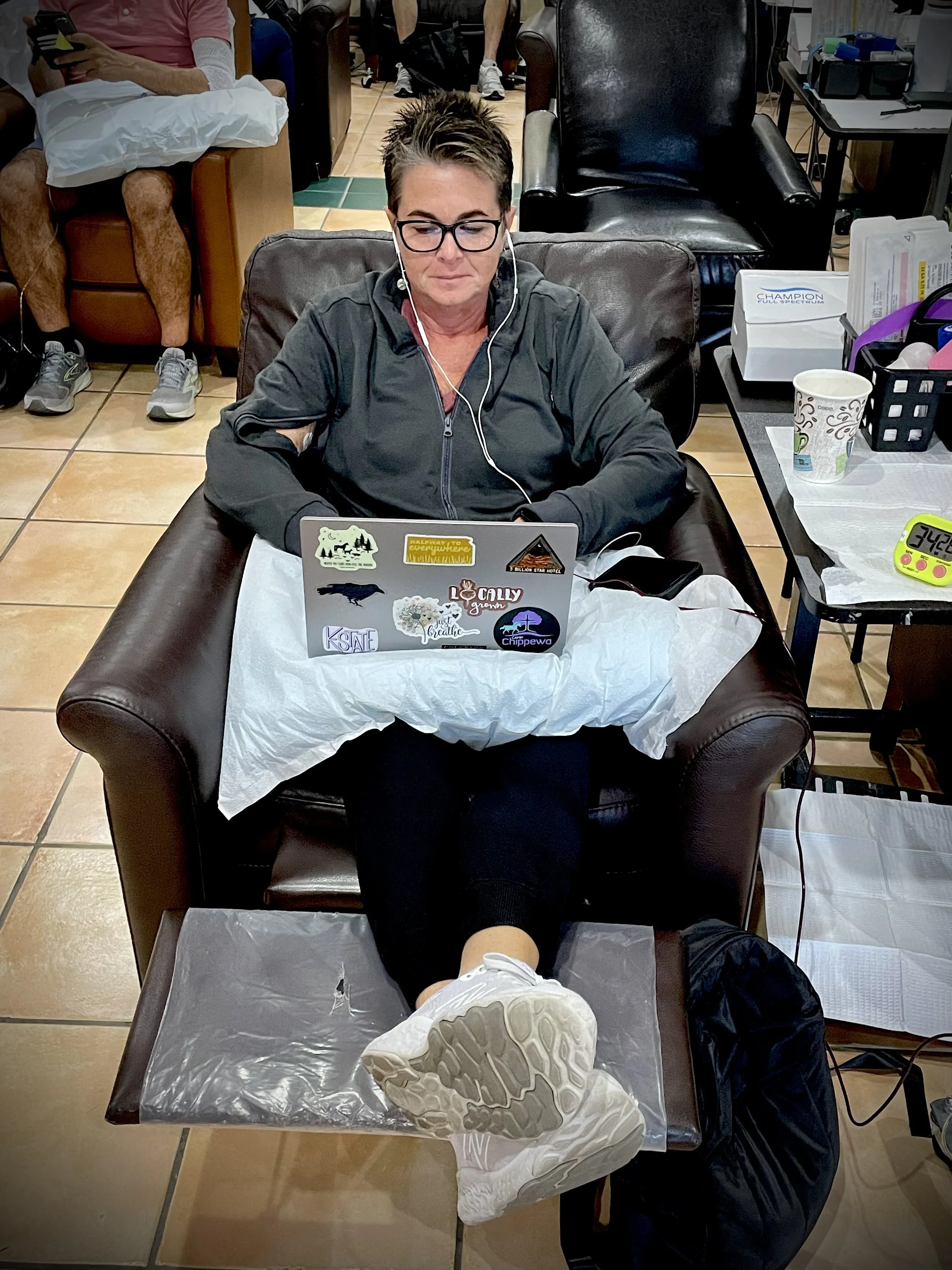 Gail with her shortest hair, glasses, and earphones sits on a leather chair, working on her laptop in the IV room. 
