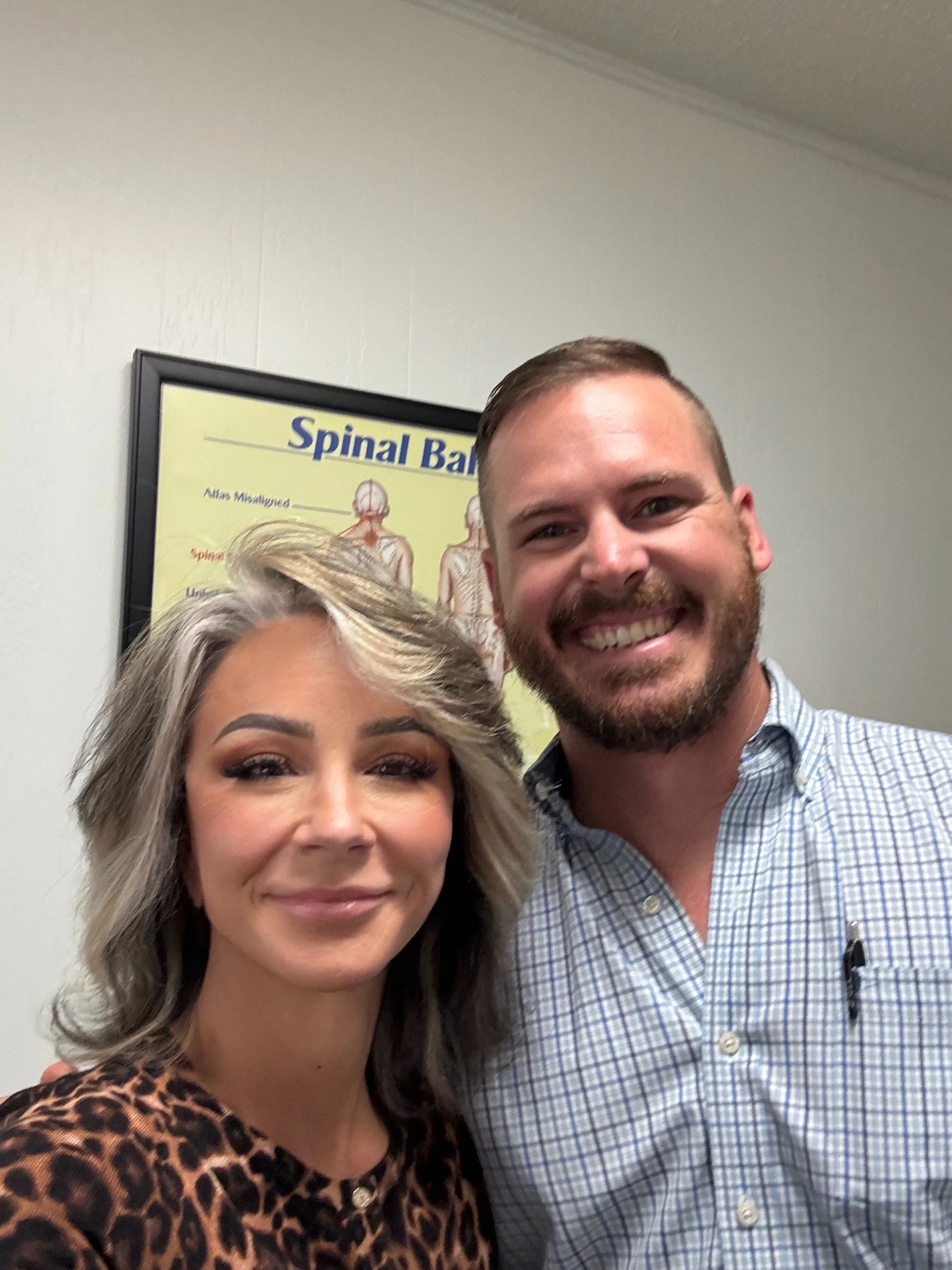 Renee DeTar and Travis from Mayer Clinic in Clearwater Florida, smiling for a selfie in front of a wall with a poster about spinal bones.