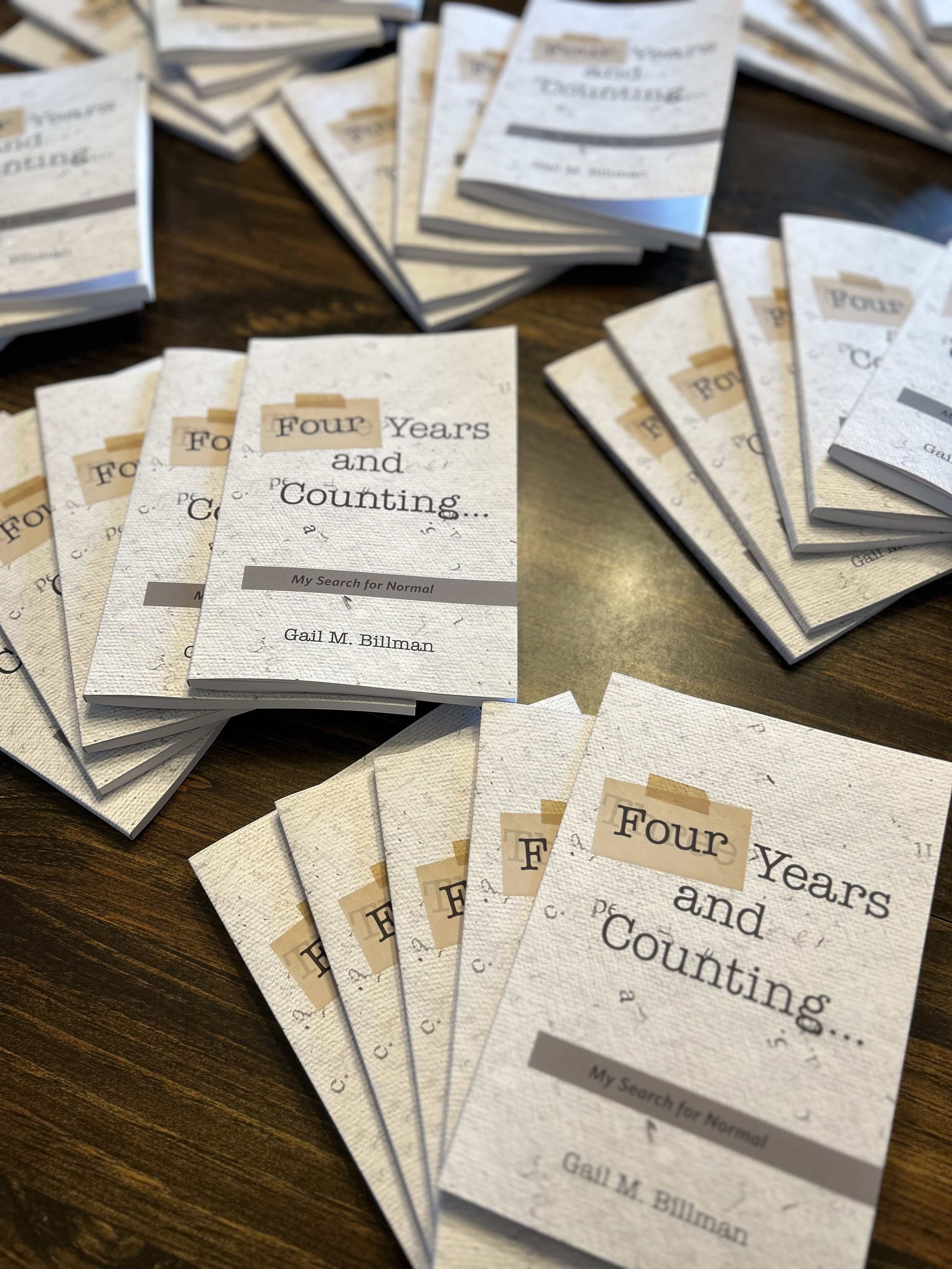 Multiple copies of a book titled 'Four Years and Counting' by Gail M. Billman are spread out on a wooden surface.