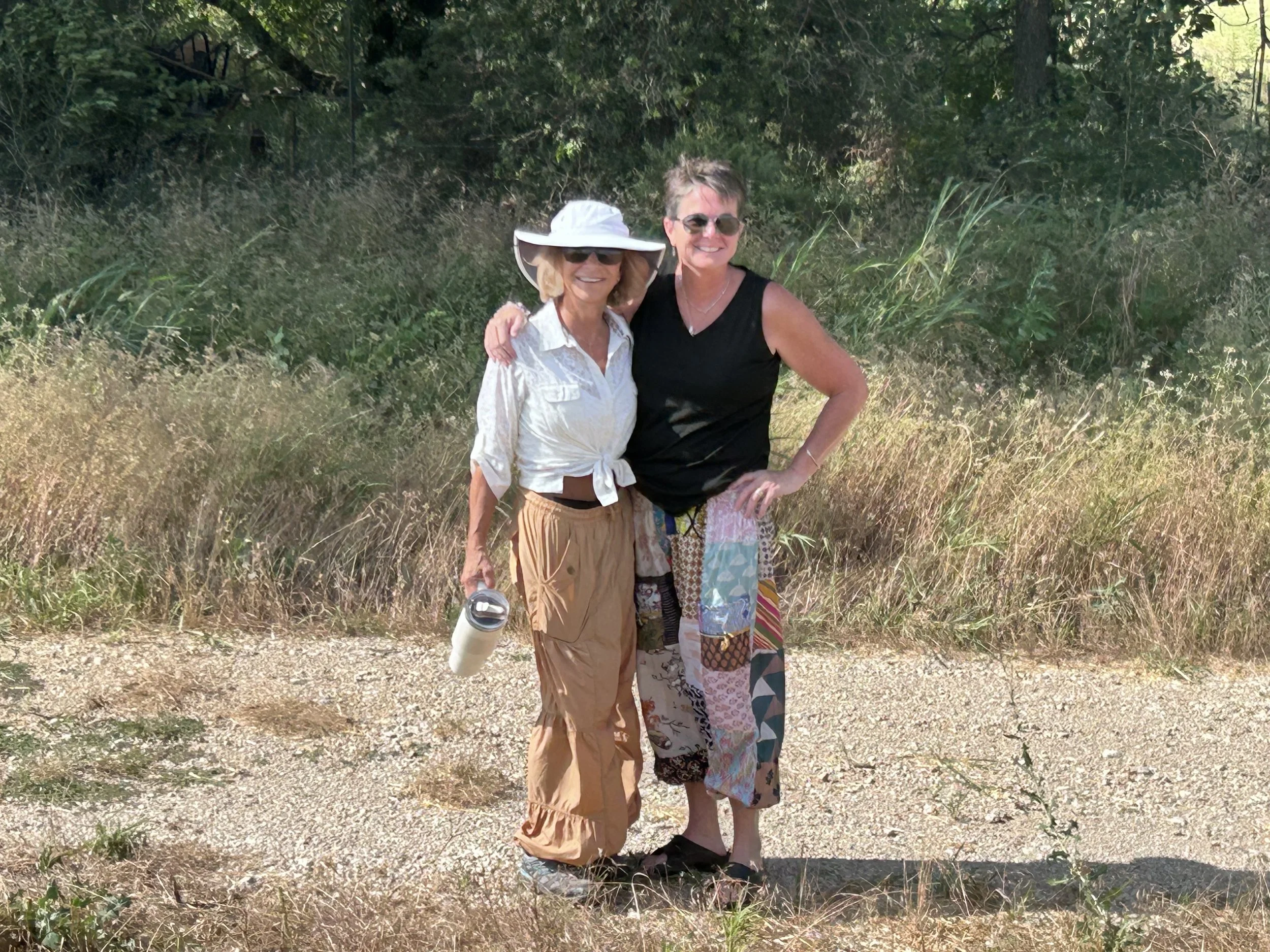 Renee DeTar and Gail Billman during harvest July 2025  standing outdoors on a dirt path surrounded by greenery, smiling, with one woman wearing a large white sun hat and sunglasses, and the other woman in a black sleeveless top and sunglasses.