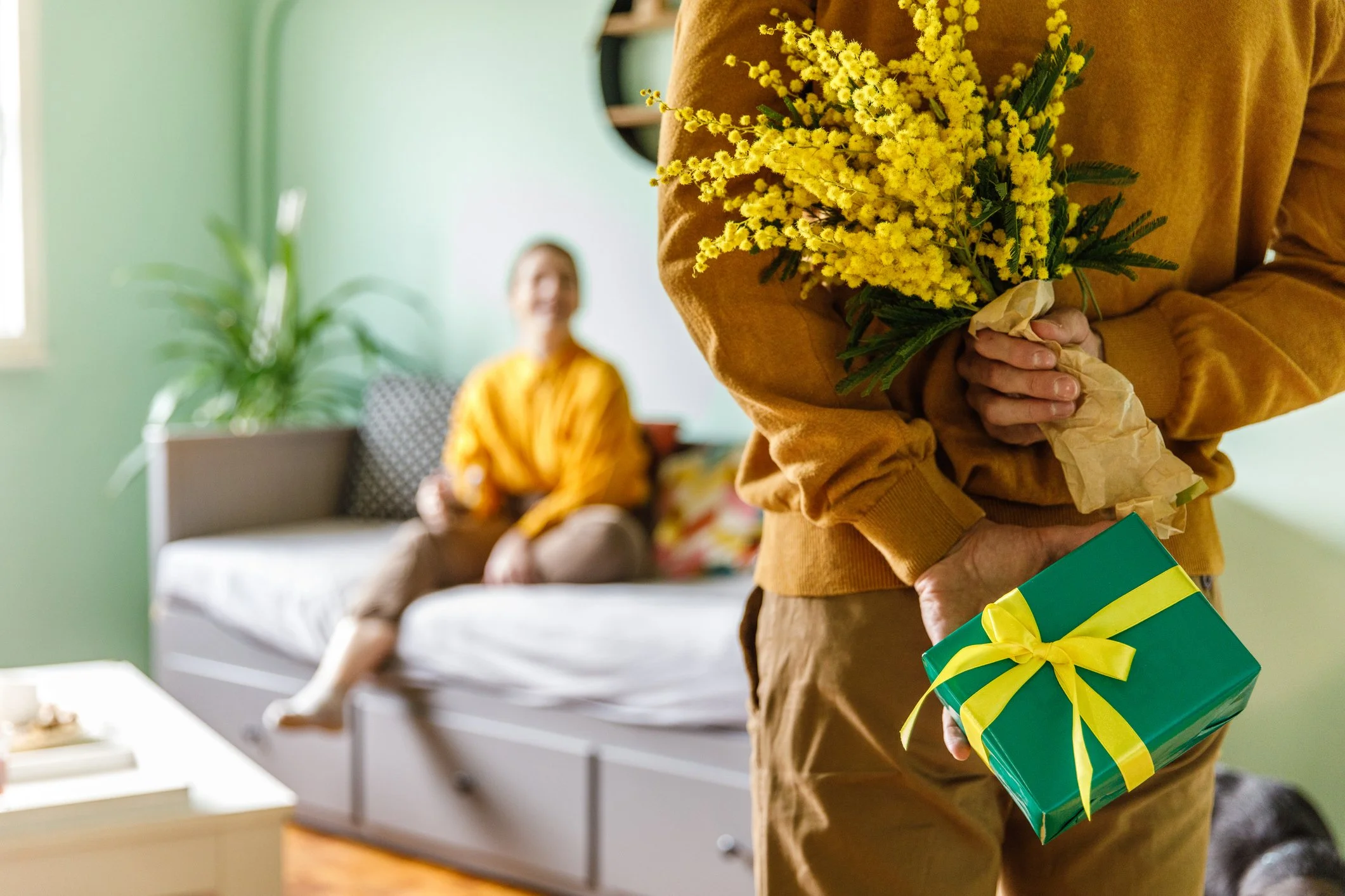 A person in a brown sweater holding a bouquet of yellow flowers and a green wrapped gift with a yellow ribbon, with another woman smiling in the background sitting on a couch.