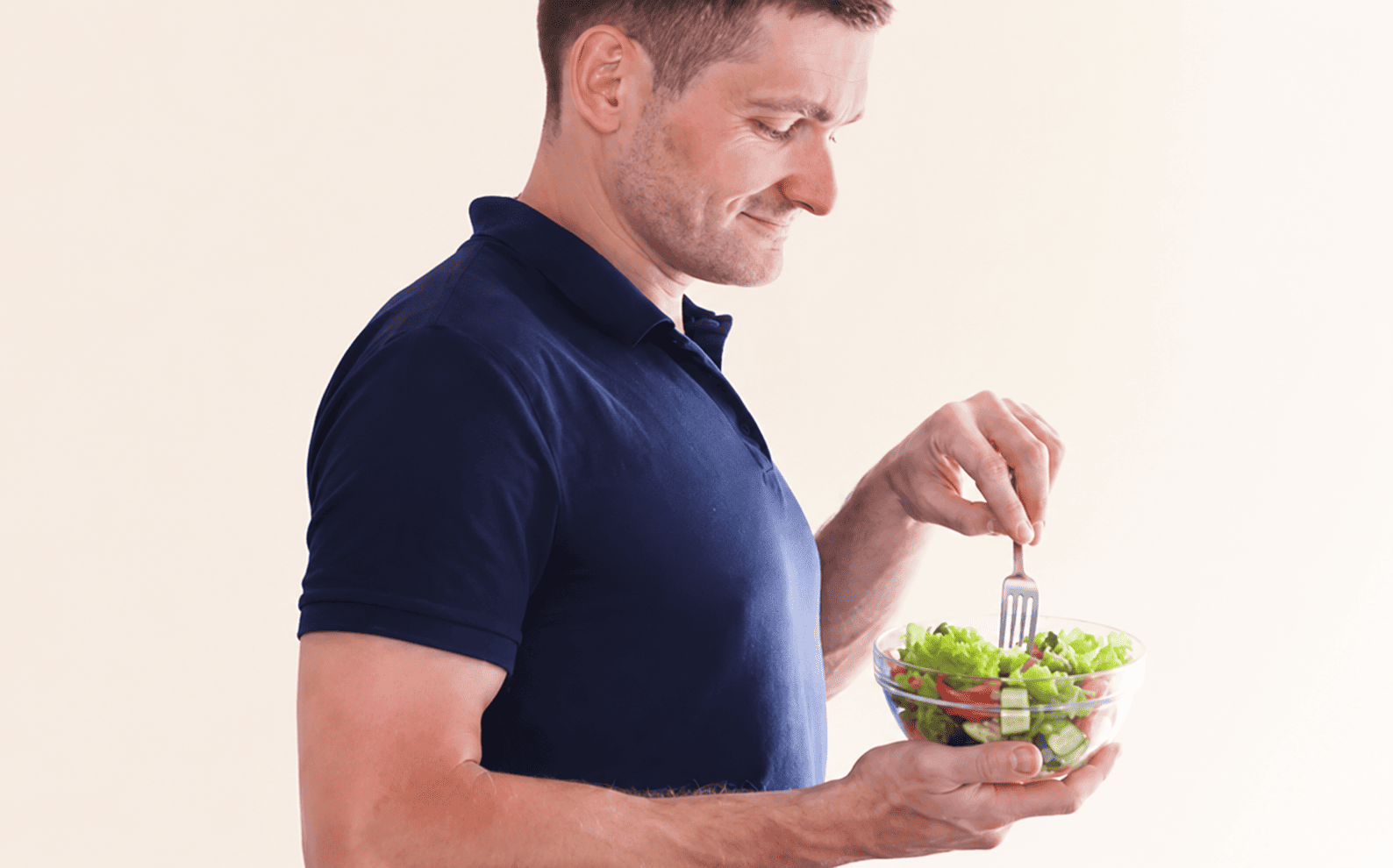 A man wearing a navy blue polo shirt holding a glass bowl of salad and eating with a fork.