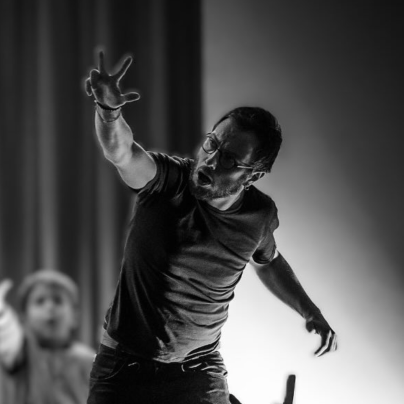 Un homme en t-shirt noir, portant des lunettes, semble crier ou parler avec intensité, avec un bras tendu vers l'avant.