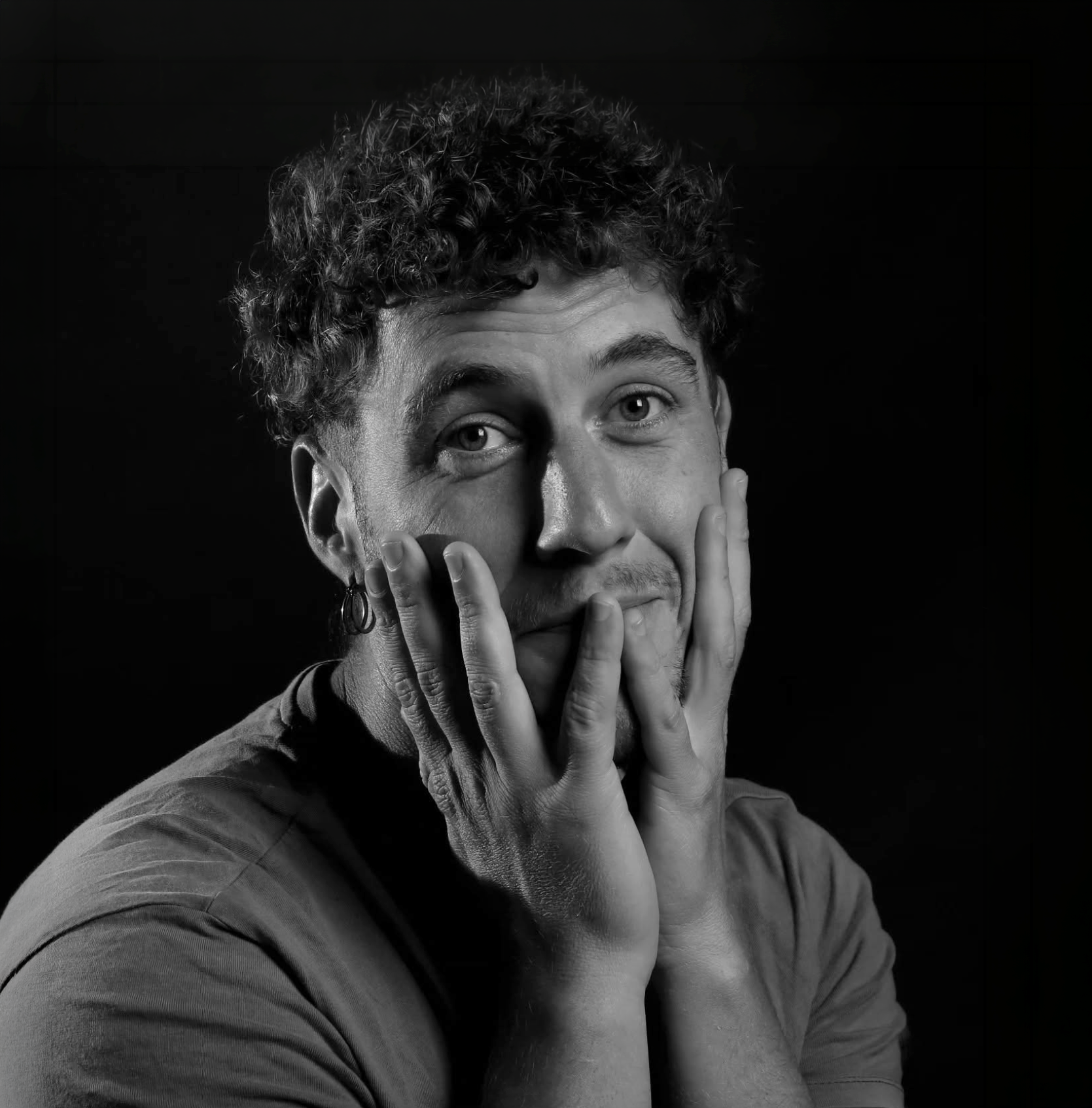 Portrait en noir et blanc d'un jeune homme avec boucles, tenant son visage avec ses mains, regardant vers l'avant.