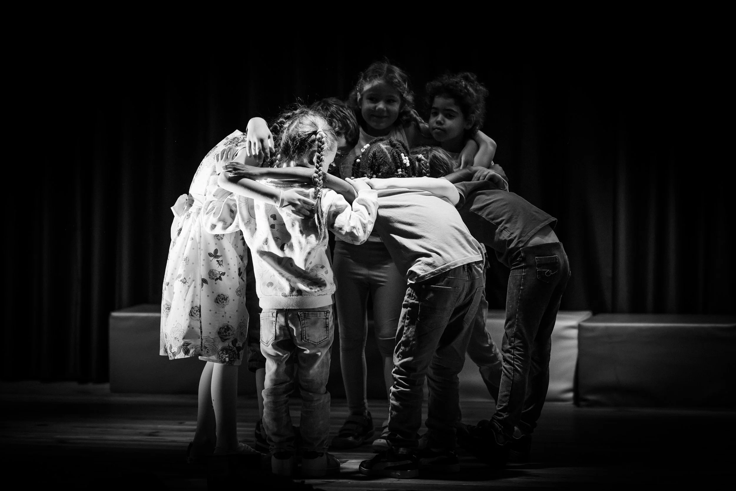 Un groupe d'enfants se rassemble en cercle sur scène, se donnant une accolade. La scène est sombre avec un fond noir et un plancher en bois. L'image est en noir et blanc.