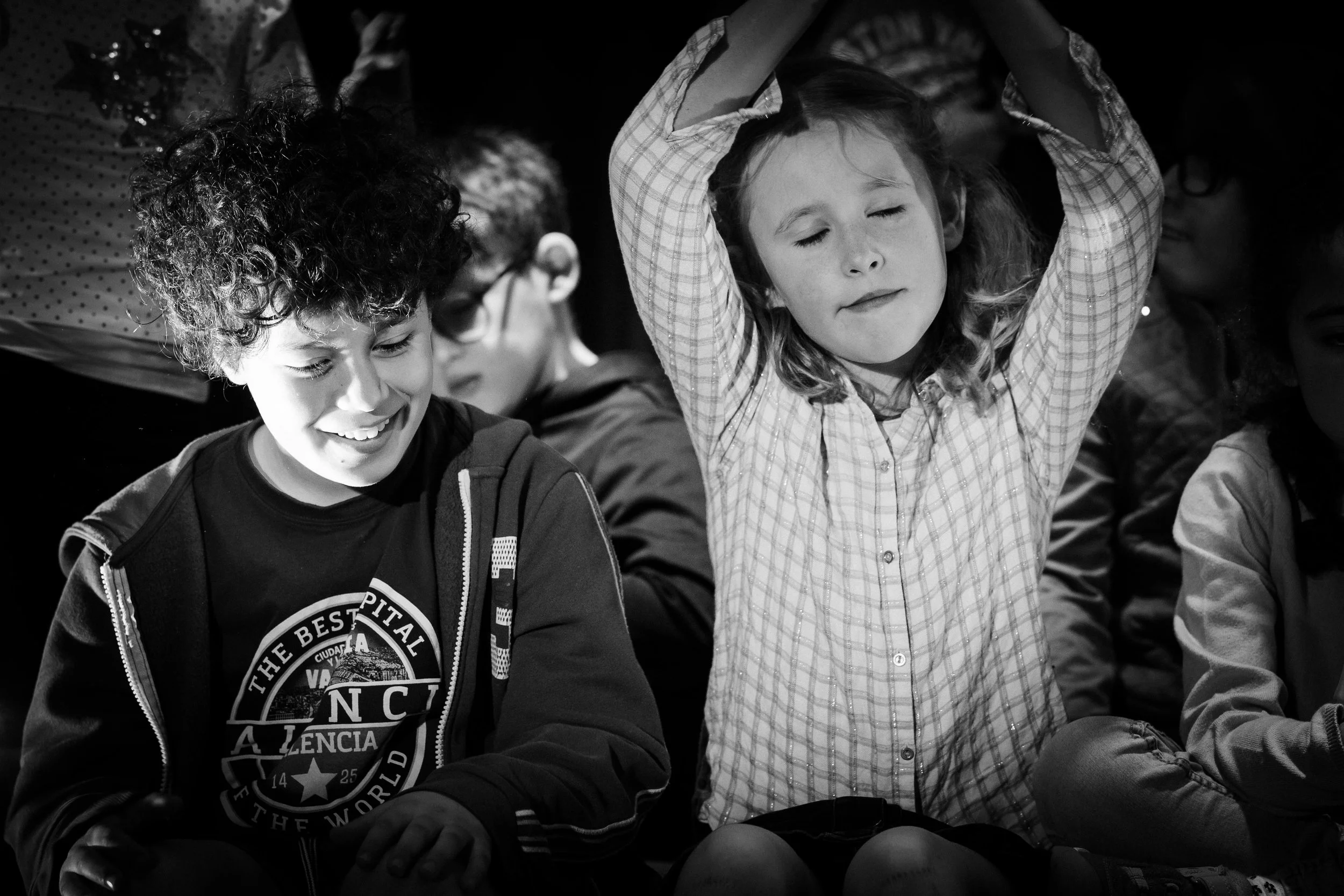 Deux enfants assis, un garçon souriant et une fille avec les yeux fermés, elle lève les bras au-dessus de sa tête.