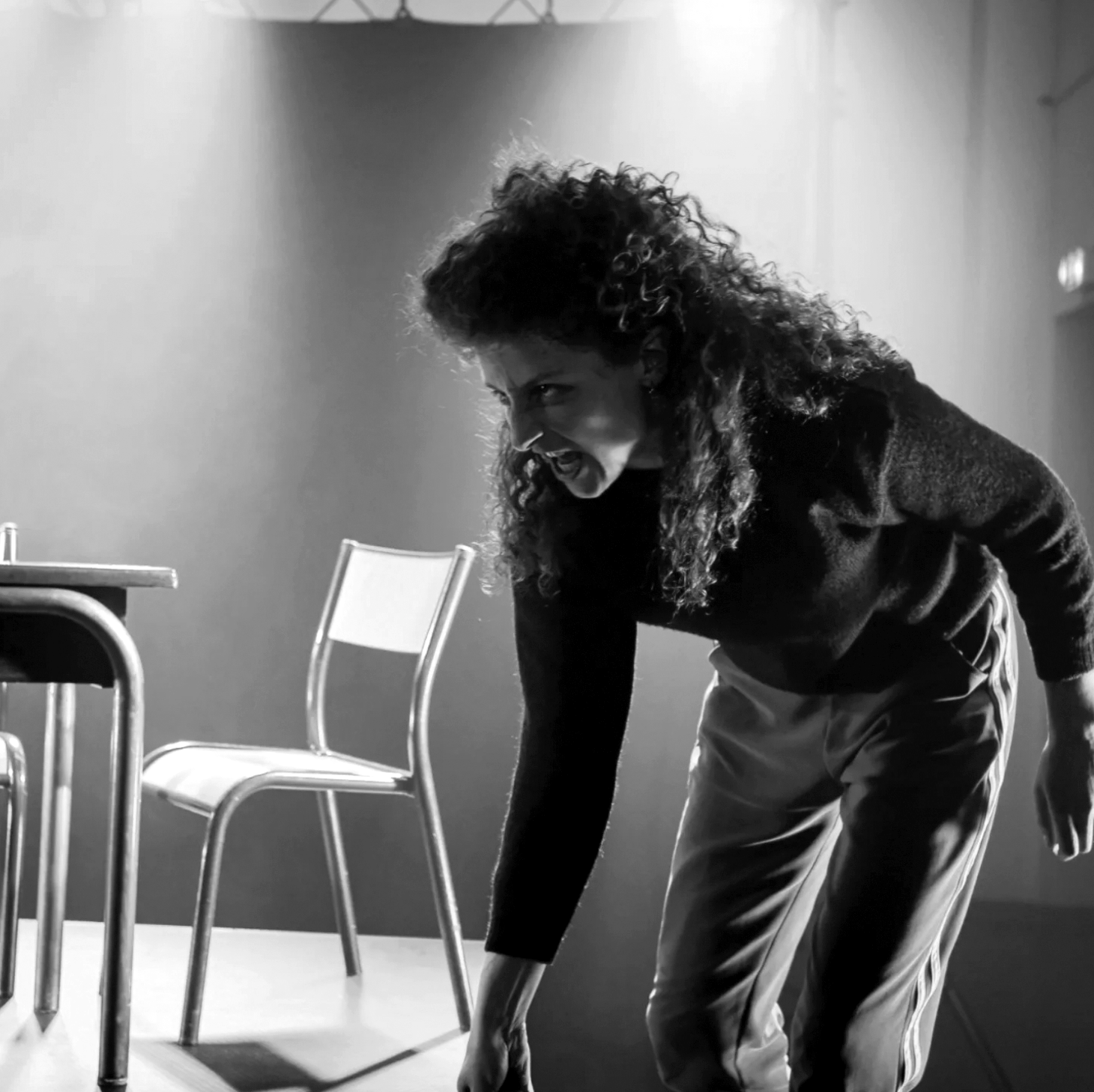 Une femme avec des cheveux bouclés, souriant de manière expressive, penchée en avant dans une pièce sombre, avec une table et une chaise à côté d'elle.