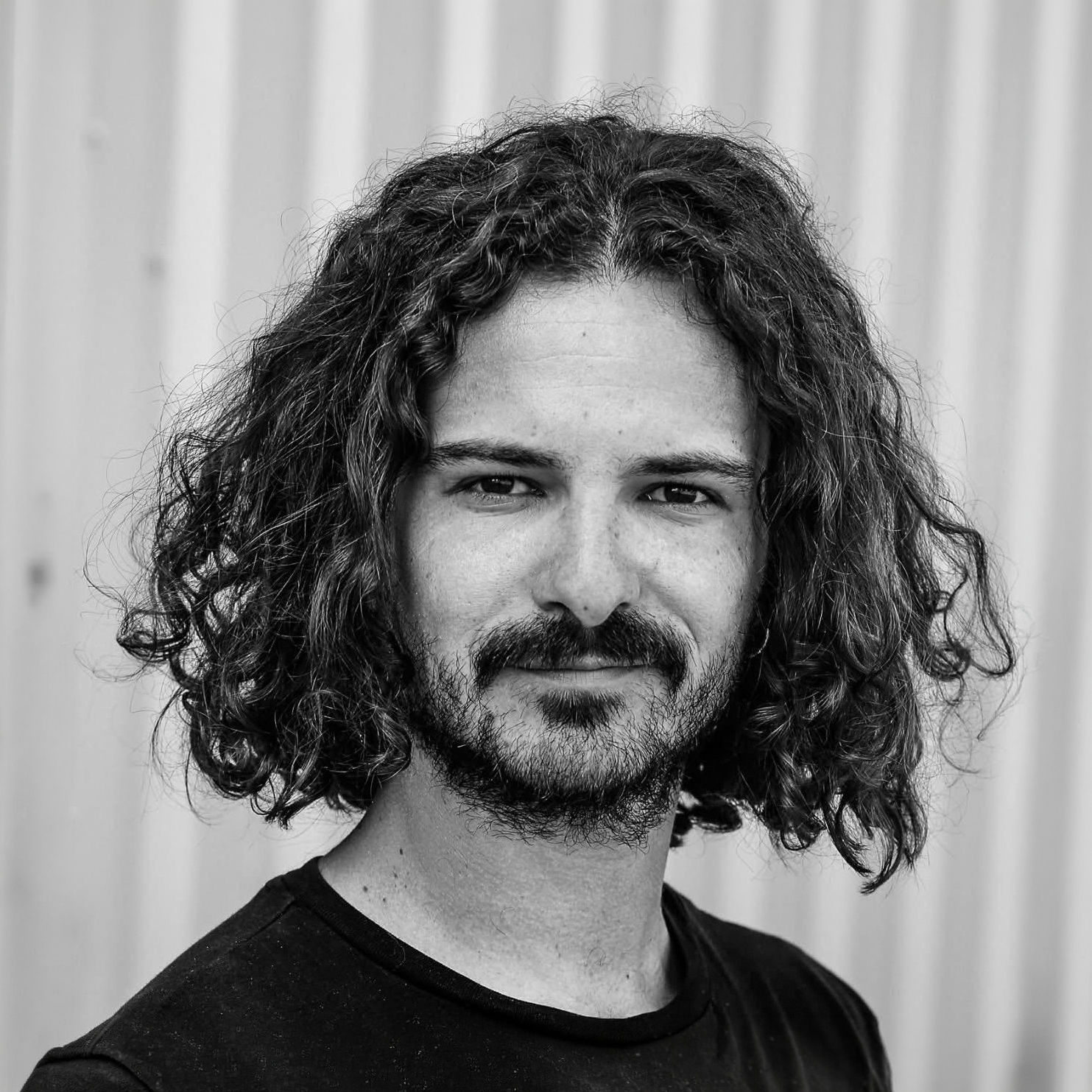 Portrait d'un homme avec des cheveux longs et bouclés, barbe et t-shirt noir, regardant vers l'appareil, fond flou.