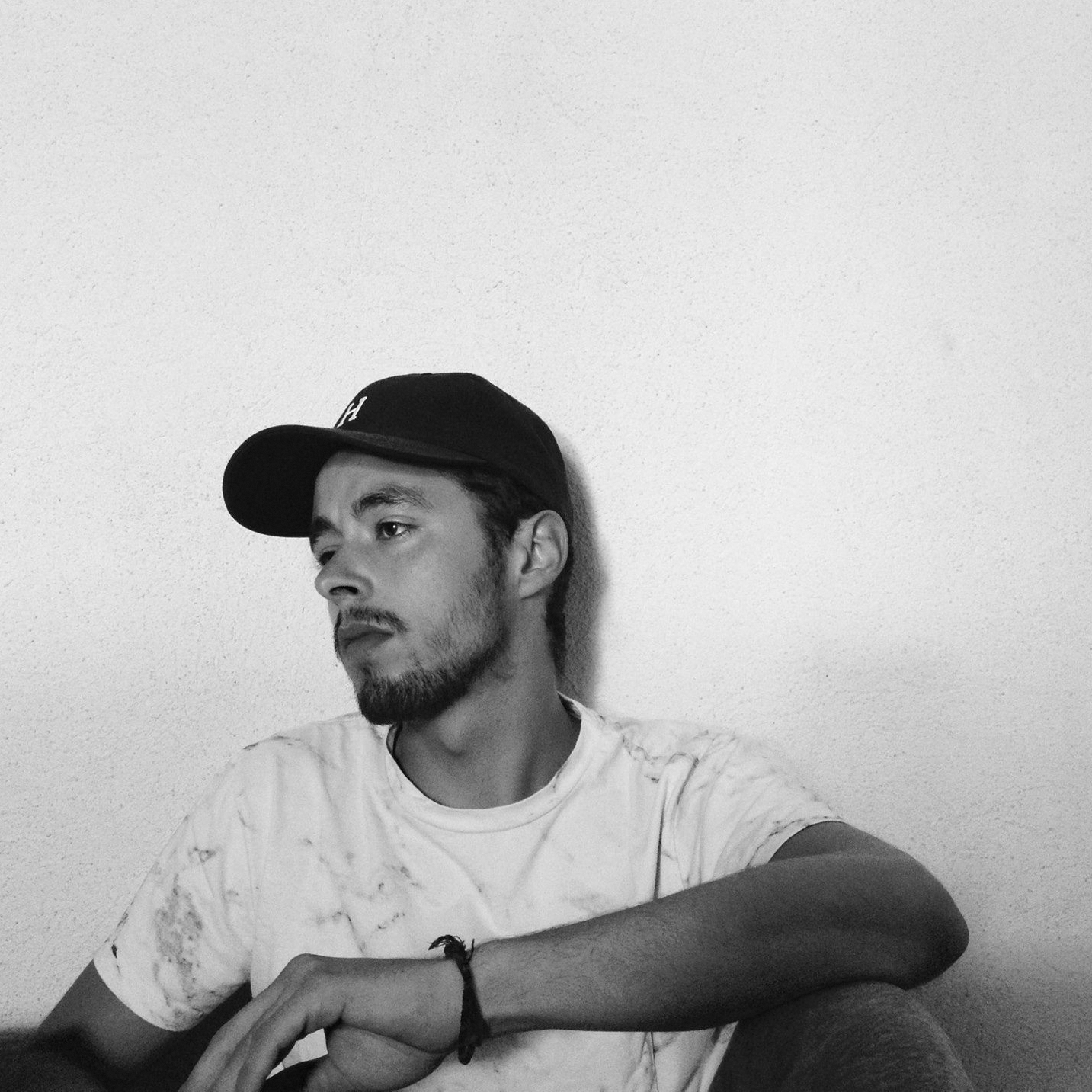 Jeune homme avec barbe portant une casquette noire et un T-shirt blanc, assis contre un mur blanc, regardant sur le côté.
