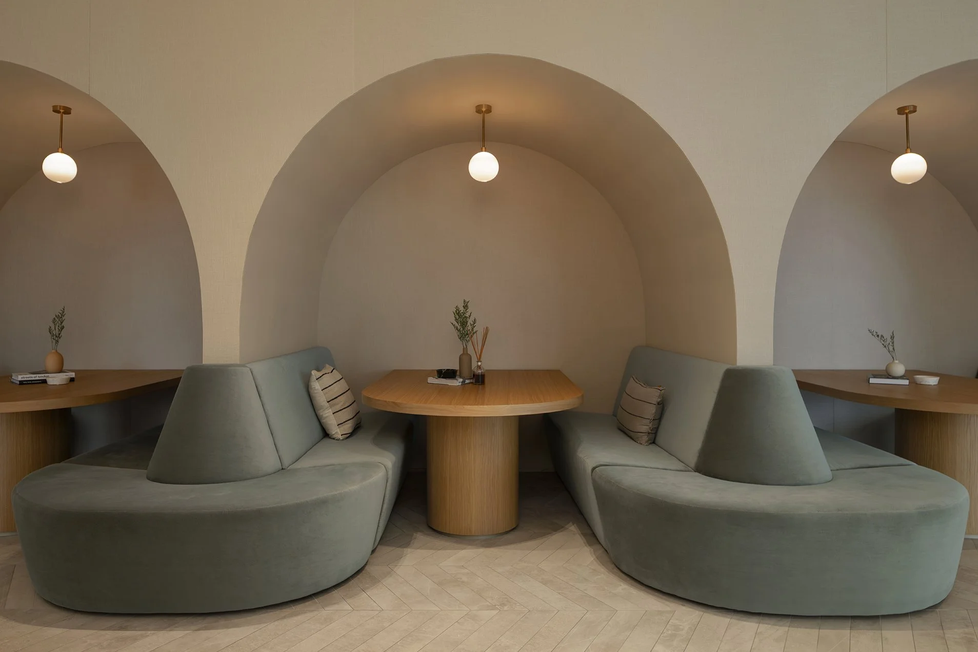 A modern interior with a circular seating area featuring a wooden table, surrounded by pastel green cushioned chairs with striped pillows. The ceiling has three semicircular arches, each with a hanging round white light. Small decorative vases with g