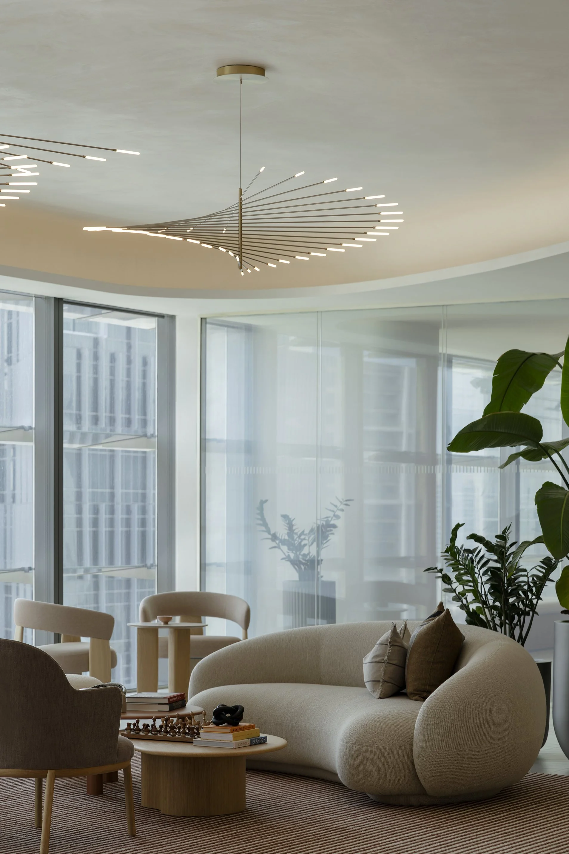 Modern living room with large windows, beige cushioned sofa, wooden side table with books and chess, brown armchair, and plants, illuminated by a contemporary chandelier.