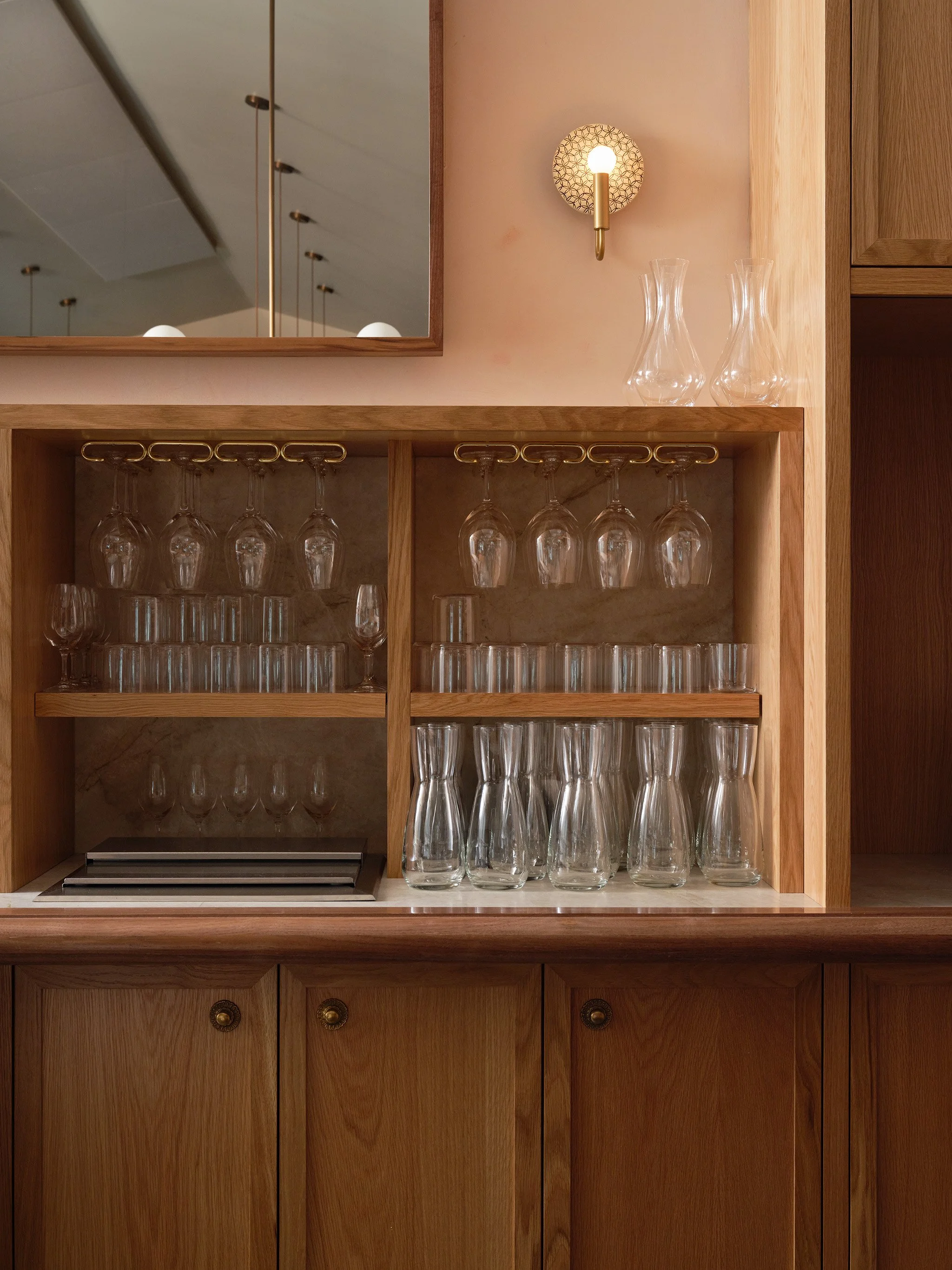 A wooden cabinet with glassware including wine glasses, water glasses, and carafes, and a mirror above it reflecting ceiling lights.