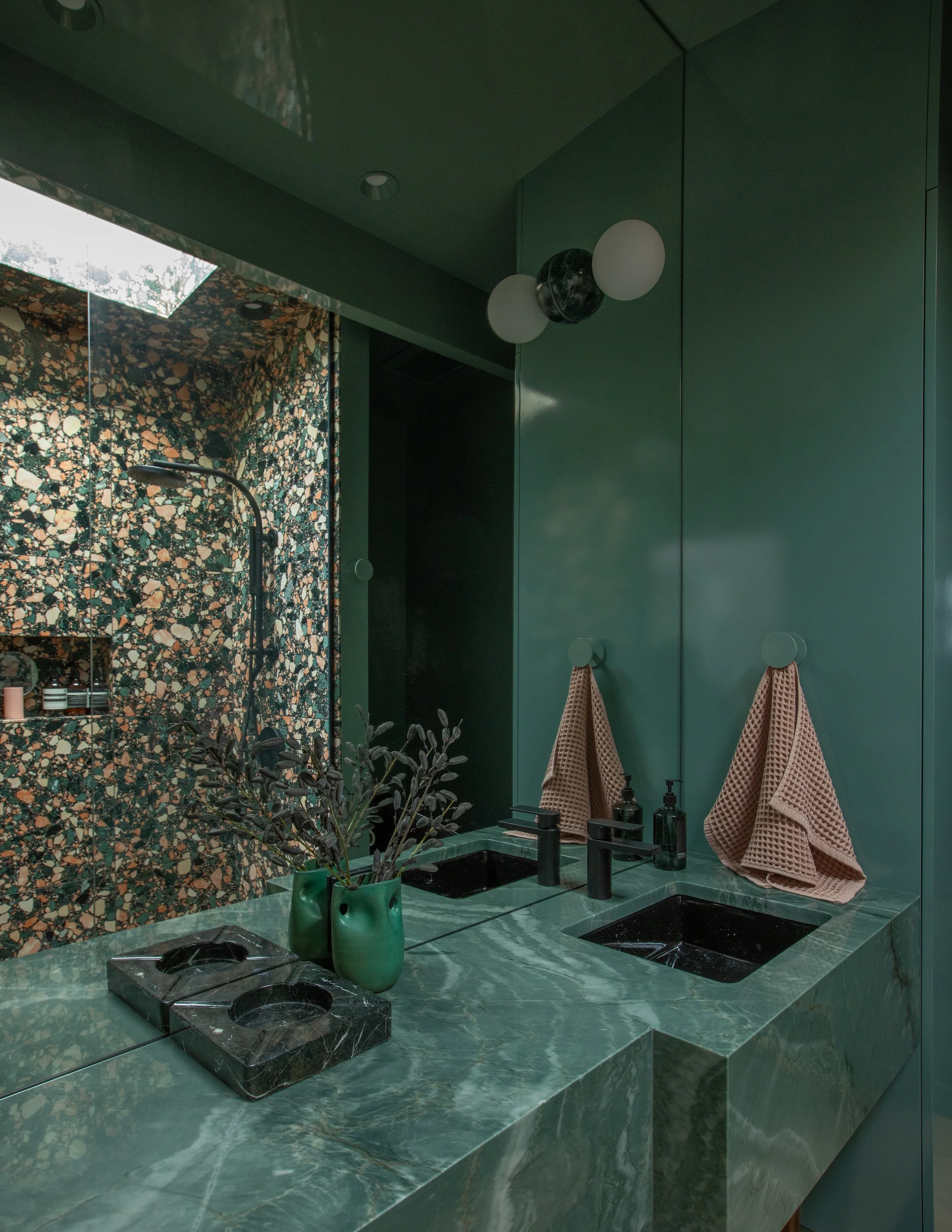 Modern bathroom with green marble countertop, black sinks, a large mirror, a potted plant, and brown towels hanging on hooks.