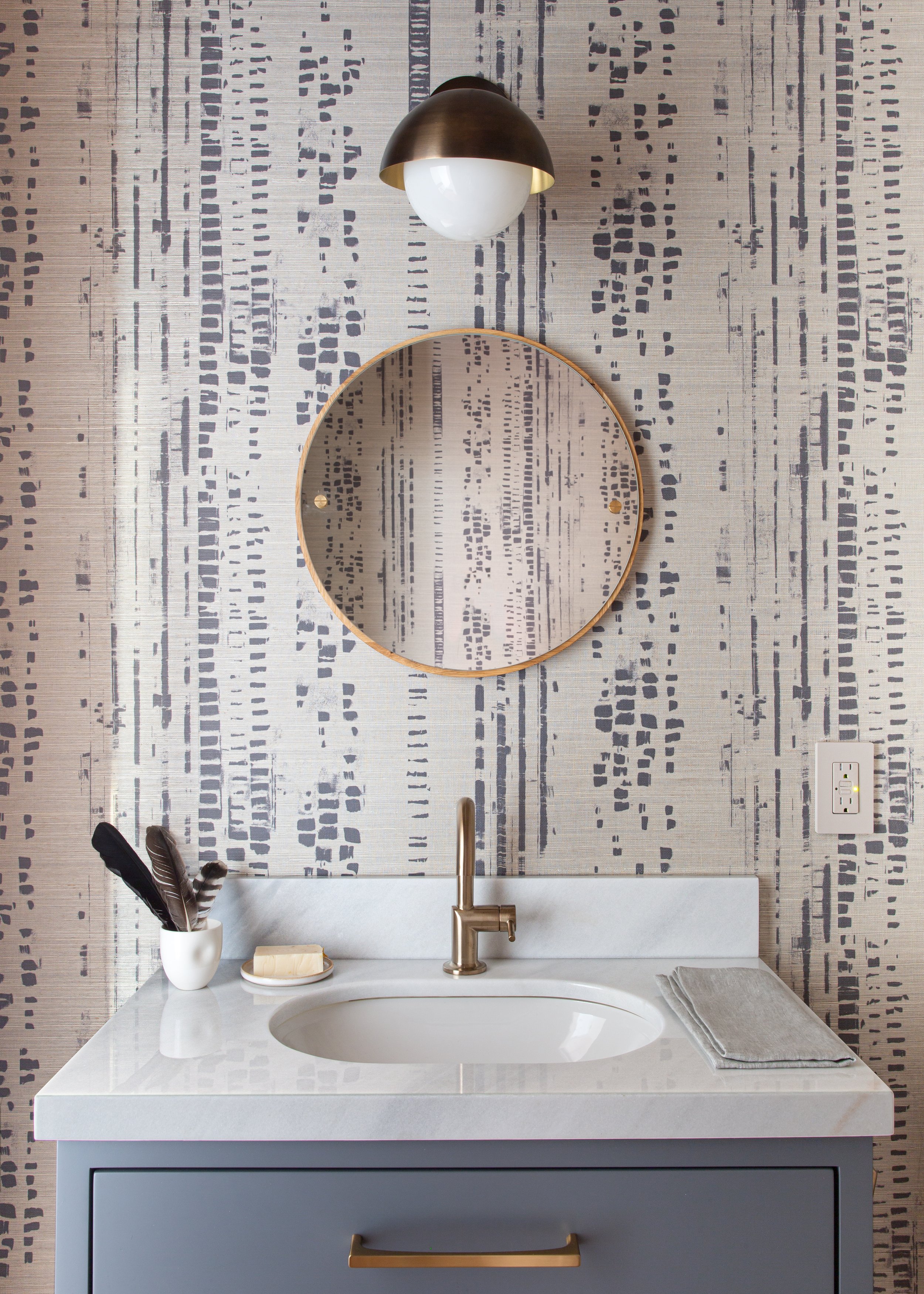 Modern bathroom with a white quartz countertop, an oval under-mount sink, a brass faucet, and a large circular mirror. The wall behind has textured wallpaper with a geometric pattern. A white vase holding feathers, a soap dish with soap, and a gray t