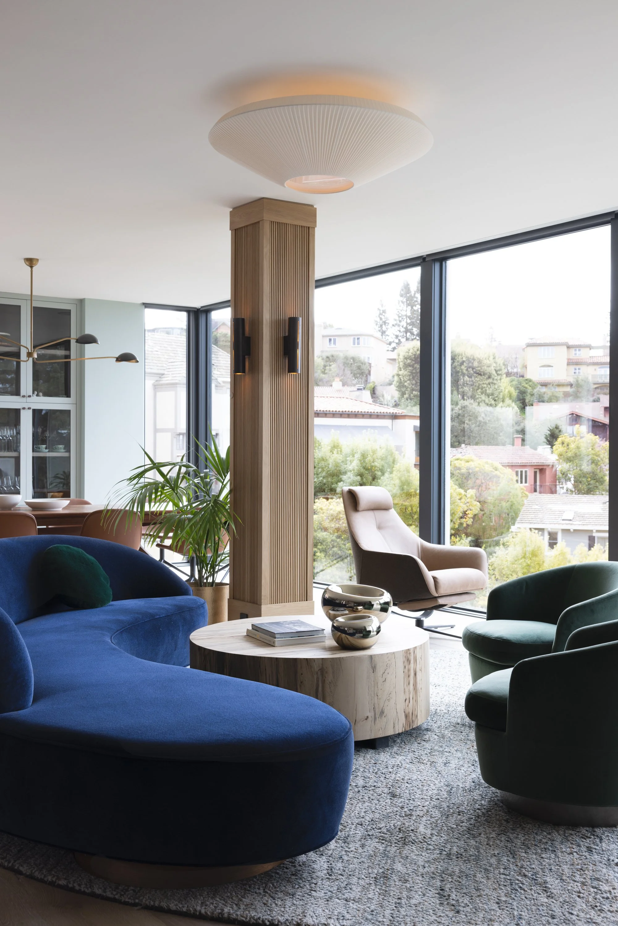 Modern living room with large windows, blue and green velvet chairs, a beige reclining chair, a round wooden coffee table with decorative bowls, and a tall wooden pillar. There is a view of a residential neighborhood with trees outside.