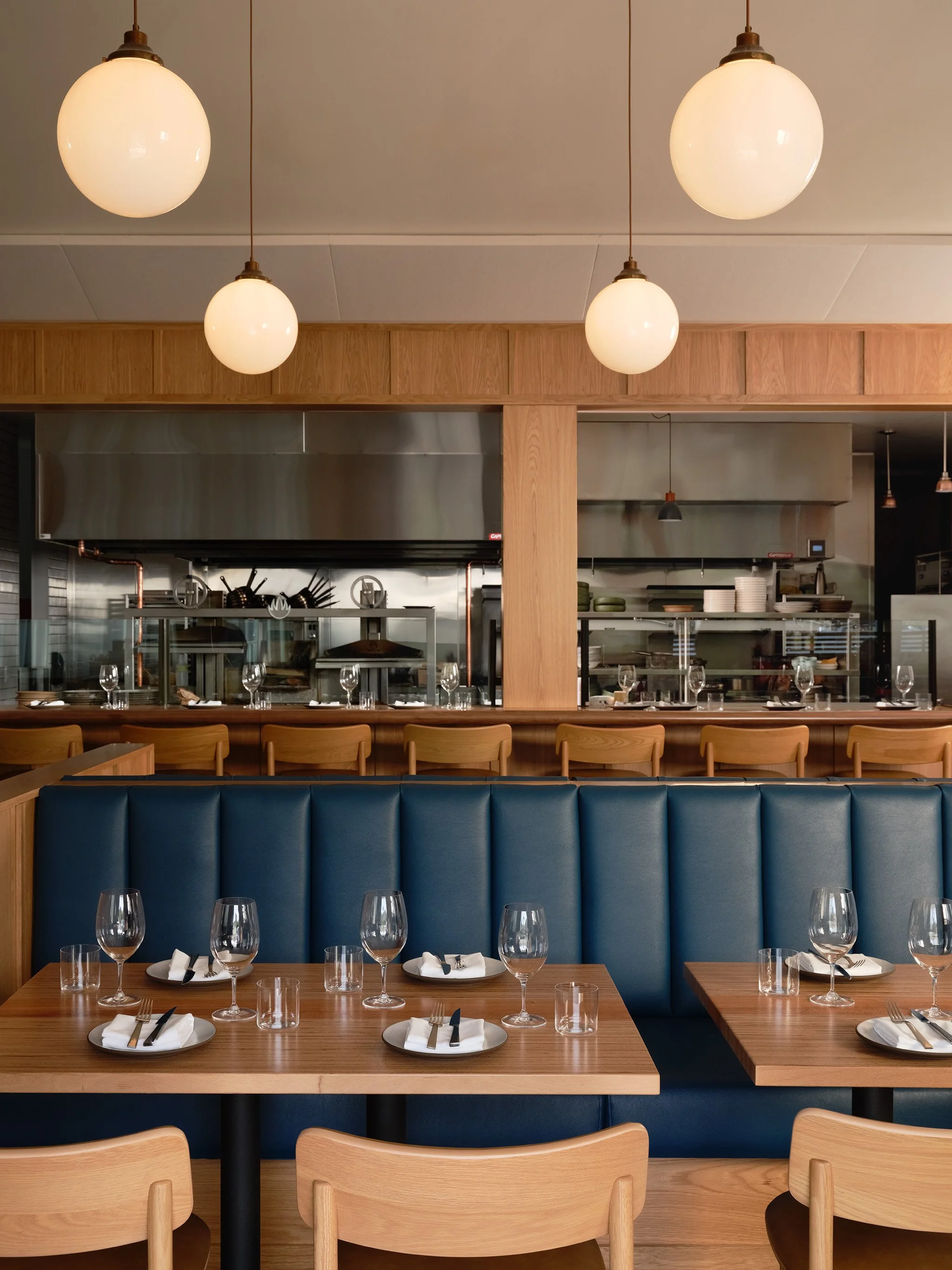 Inside of a restaurant with a wooden dining counter, blue upholstered seating, and glassware and silverware set on tables. Large hanging globe lights illuminate the space, with an open kitchen visible in the background.