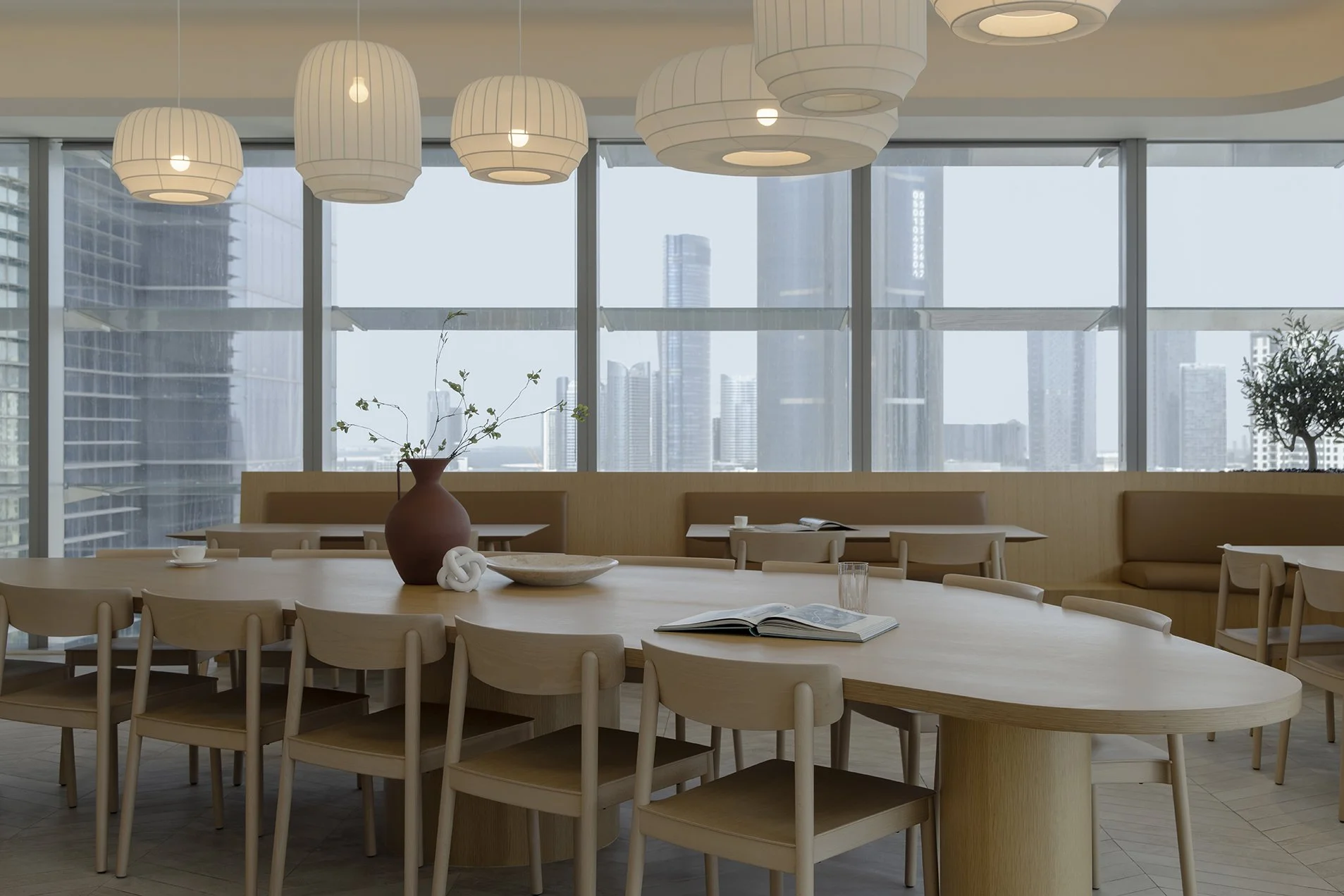 Modern dining area with beige chairs around a large oval wooden table, decorated with a brown vase, white knot sculpture, bowls, cups, and an open magazine. Large windows with a city skyline in the background, and hanging white pendant lights above.
