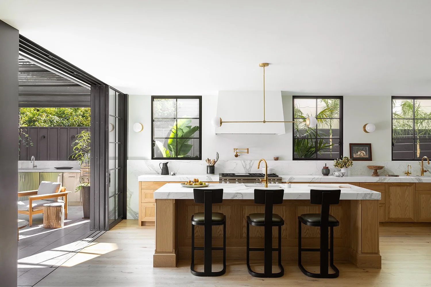 Modern kitchen with marble countertops, wooden cabinets, black bar stools, pendant light, and large windows with greenery outside.