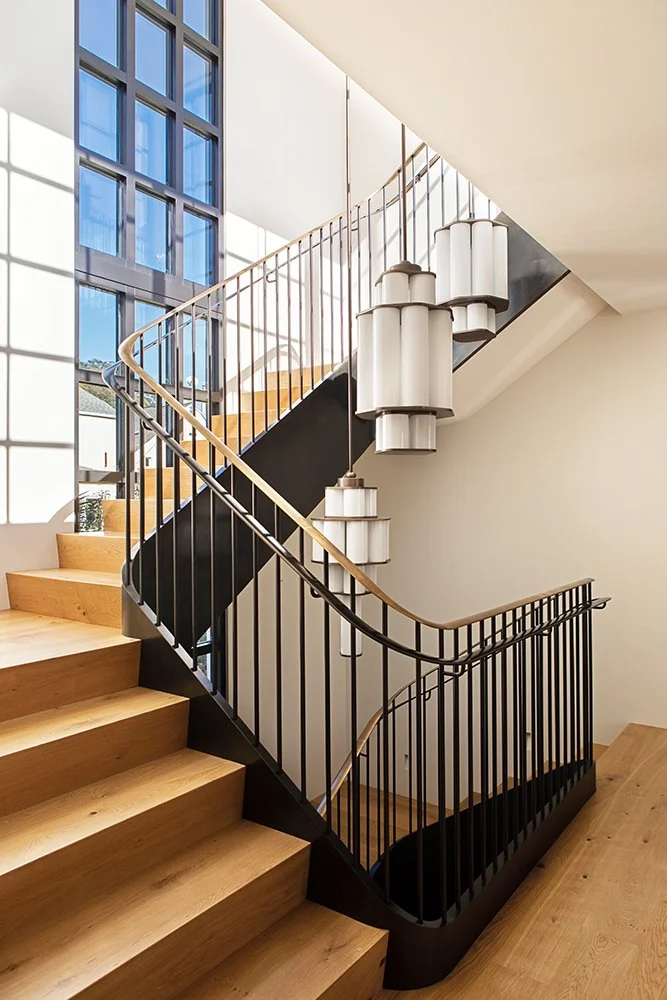 Interior staircase with wooden steps, black metal railings, and hanging cylindrical light fixtures, large windows with blue sky reflections, and minimalistic modern design.