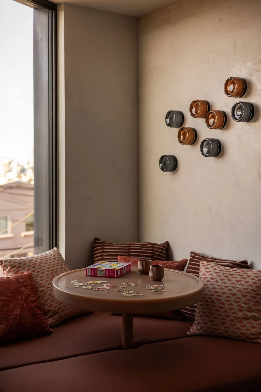 A cozy corner with a low table surrounded by pillows, puzzle pieces, and two cups on the table. A window shows an outside cityscape, and decorative wall-mounted cups are on the beige wall.