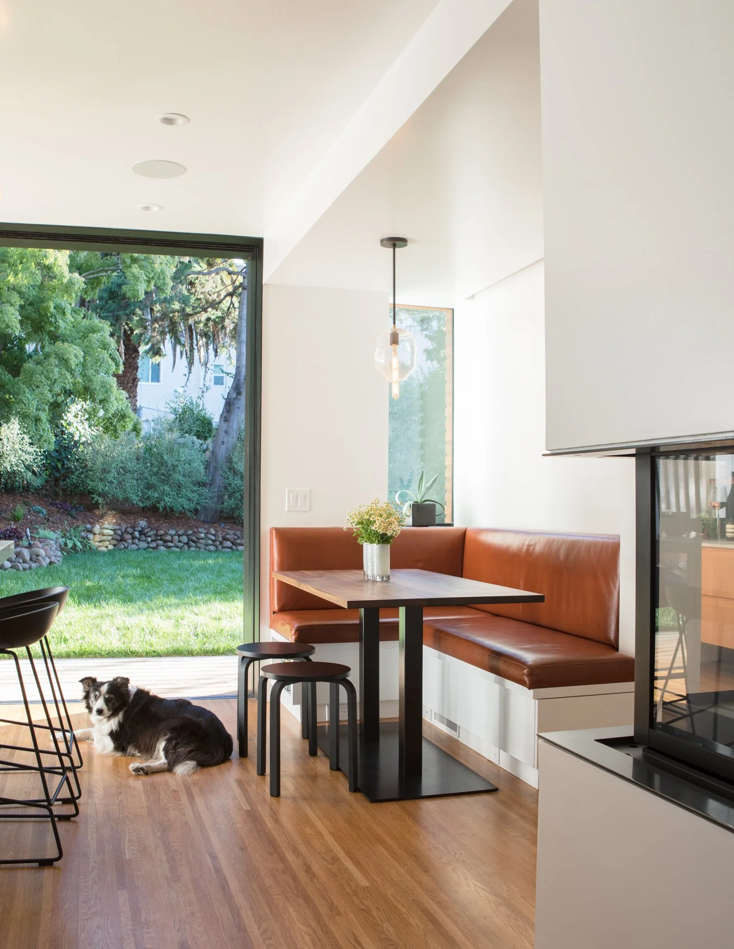 Modern kitchen or dining area with a large sliding glass door leading to a backyard with trees and grass. Inside is a corner bench seat with brown leather cushions, a wooden table with a white vase of yellow flowers, and black bar stools. A small bla