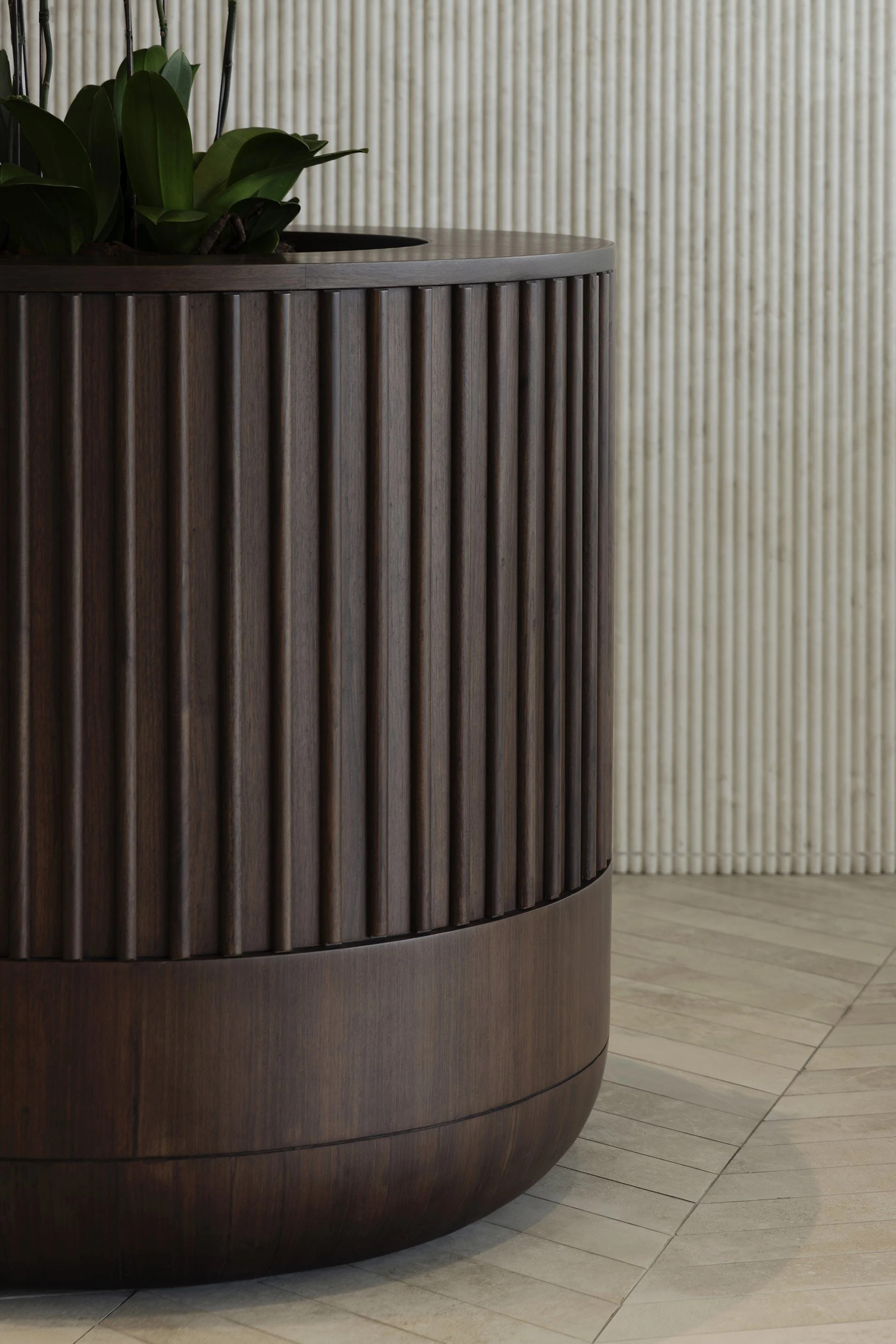 A round wooden planter with green plants inside, placed on a tiled floor with a beige textured wall in the background.