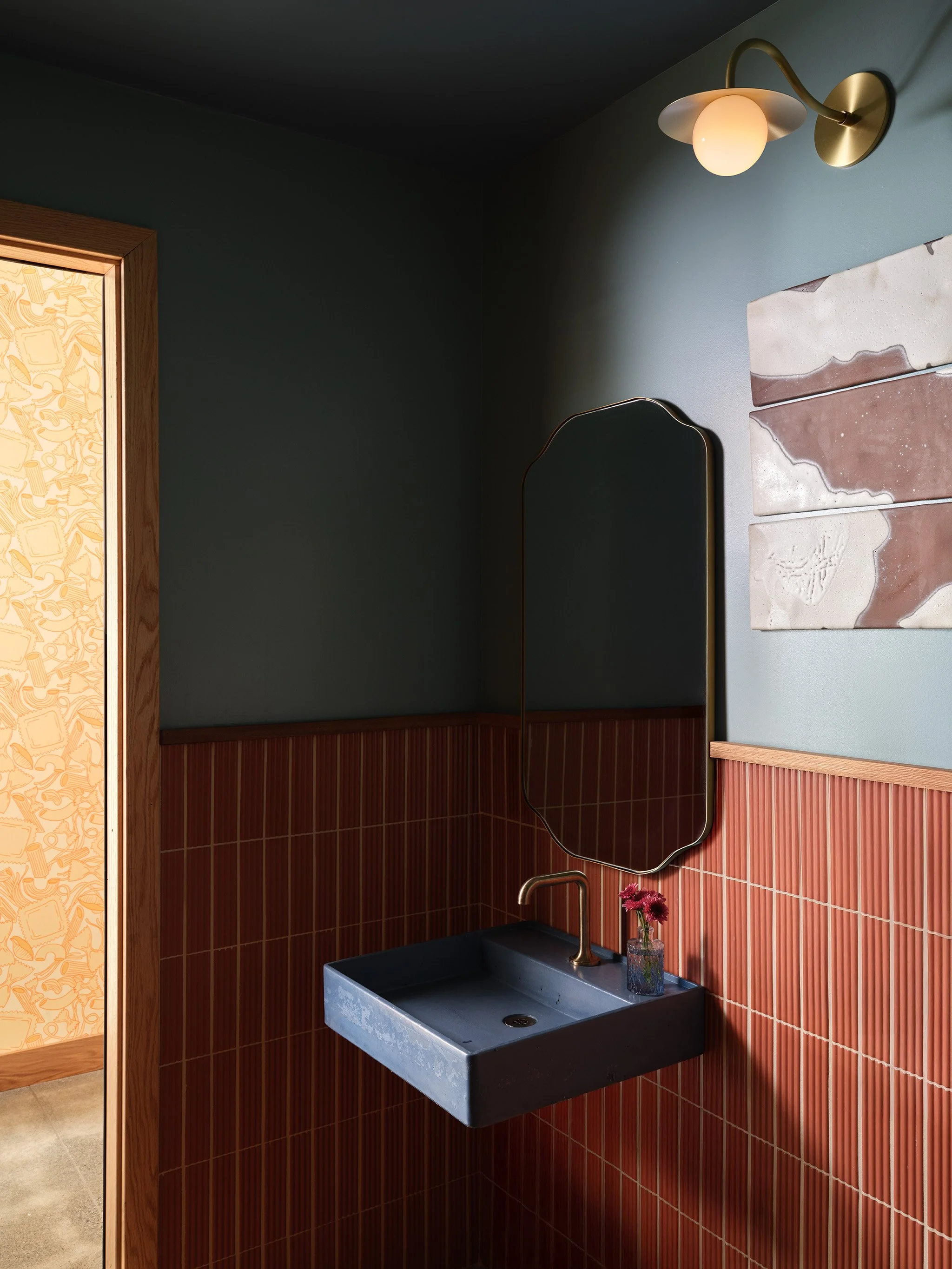 Interior of a room with a wall-mounted black sink with gold faucet, a rectangular mirror with a gold frame, white and pink abstract art on the wall, a brass wall sconce with a round light bulb, and a small vase with pink flowers on the sink.