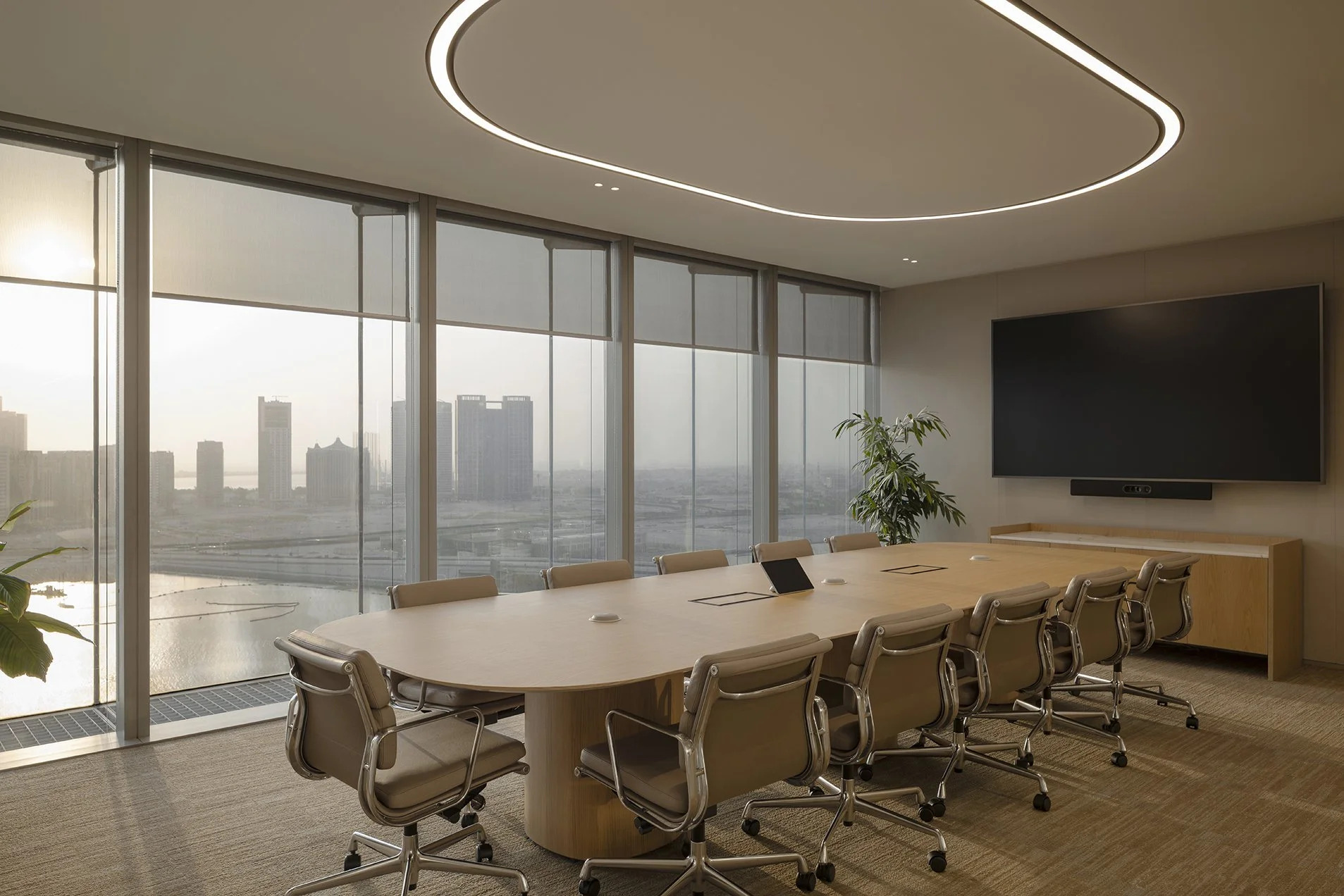 Modern conference room with a large oval table and beige office chairs, large windows with city view, a wall-mounted flat-screen TV, and a circuit design ceiling light.