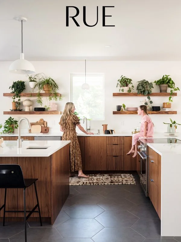 A modern kitchen with two women, one standing at the counter and the other sitting on the stove, surrounded by plants on wooden shelves, and a window in the background. The word 'RUE' is displayed at the top.