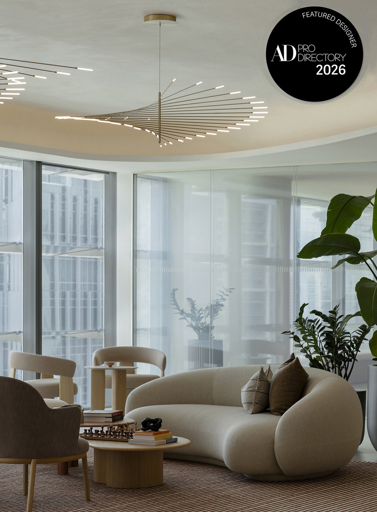 Modern living room with curved beige sofa, armchairs, wooden coffee table, and plants. Floor-to-ceiling windows with city views and contemporary lighting fixture on ceiling.