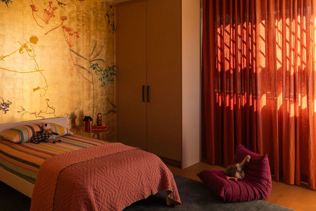 A cozy bedroom with a bed covered in a striped quilt and two pillows, a stuffed animal on the bed, a side table with a lamp and ukulele, a large wardrobe, and orange curtains with sunlight casting shadows inside the room, a small stuffed animal on a 