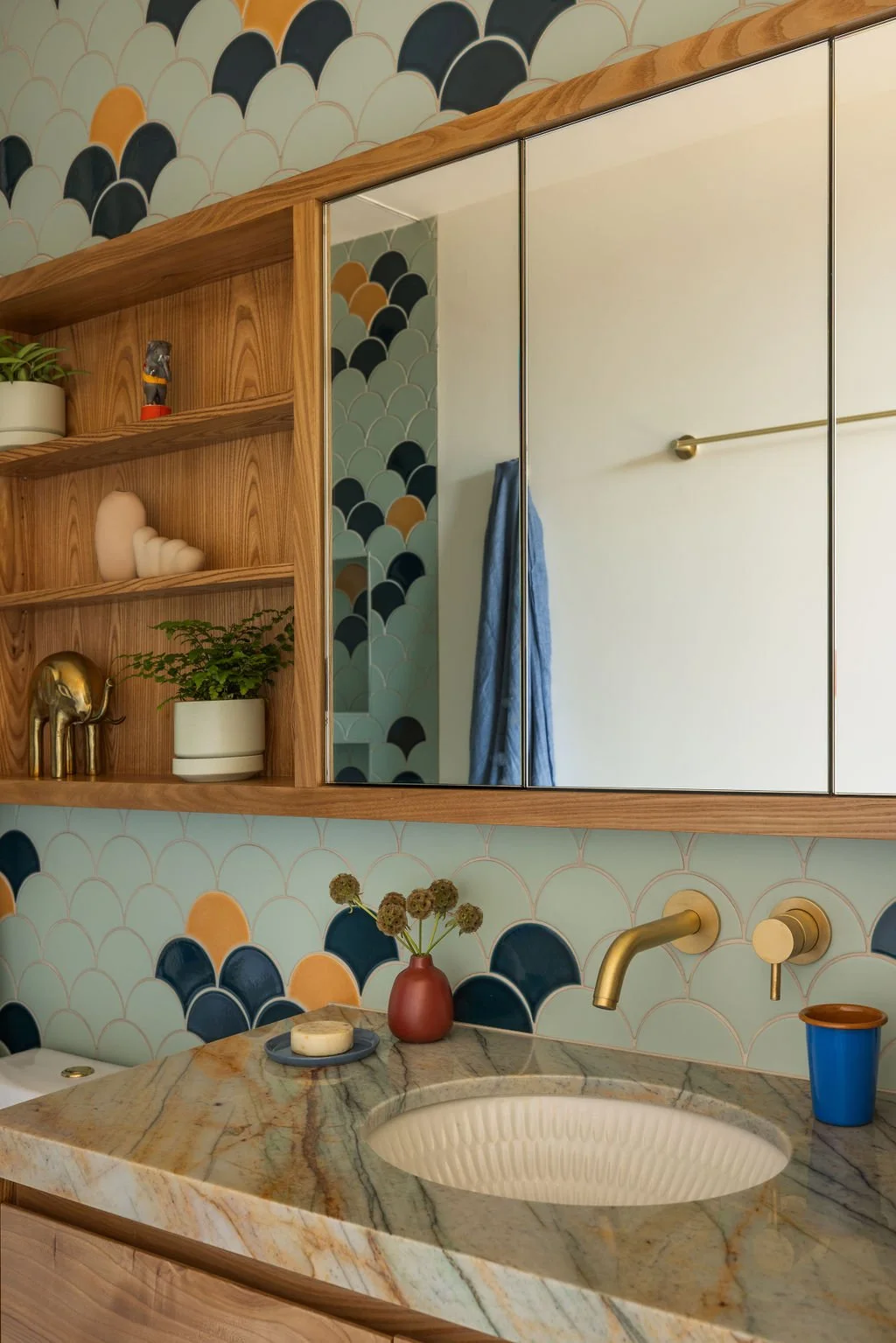 A bathroom with a green, beige, and black scallop-patterned wallpaper, a marble countertop with an integrated sink, a gold faucet, and a gold soap dispenser. There are decorative items on a wooden shelf, including a potted plant, a small sculpture, a