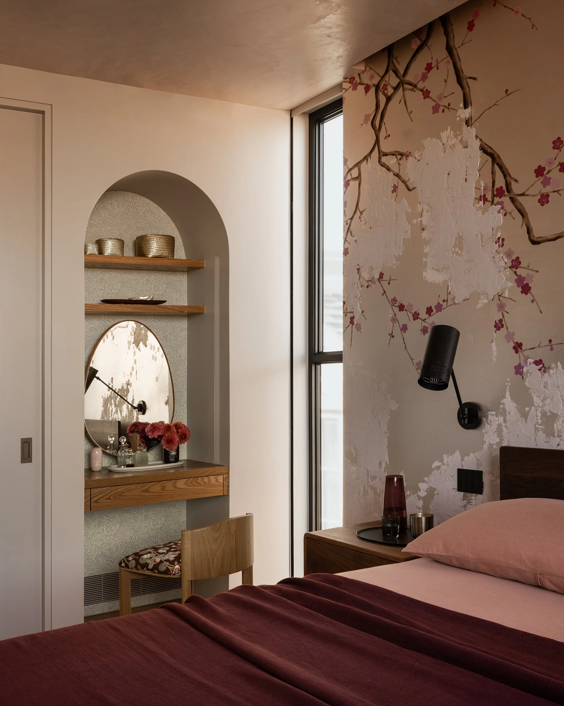 Contemporary bedroom with a bed with a maroon bedspread, side table with glassware and red vase, a wooden chair with patterned cushion, wall decor with cherry blossom mural, built-in shelves with decorative items, and a medical or reading lamp.