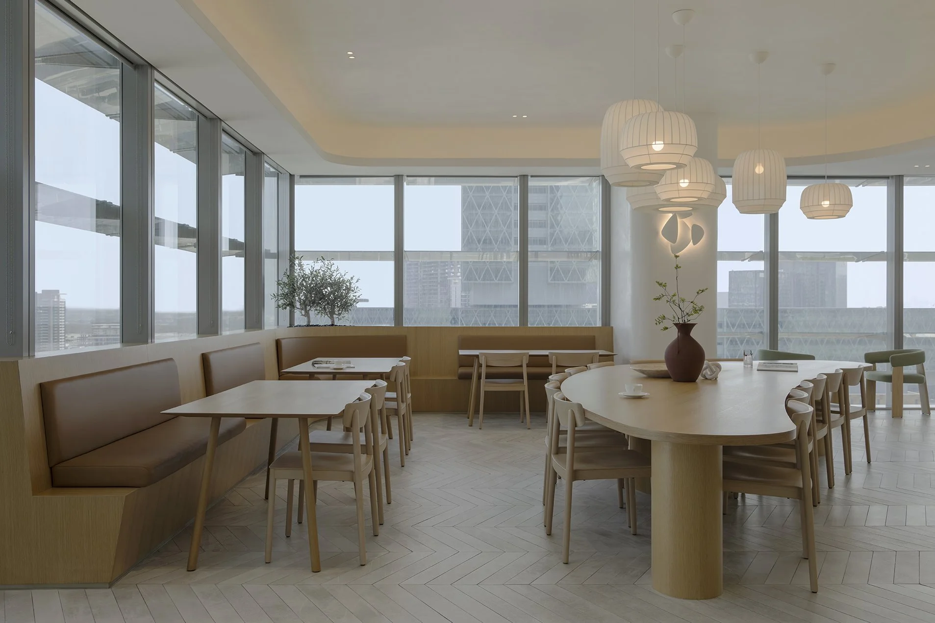 Modern restaurant or cafe interior with large floor-to-ceiling windows, round and rectangular wooden tables, beige and light green chairs, a wooden bench seating area, ceiling pendant lights, potted plants, and a cityscape view outside.