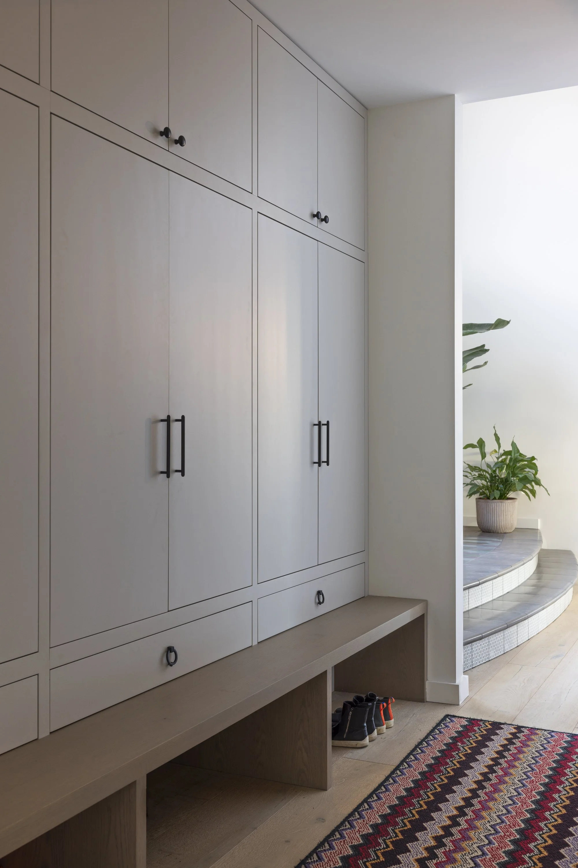 A close-up of white built-in cabinets with black handles in a modern interior, with a plant in a pot visible in the background on a curved white tiled surface.