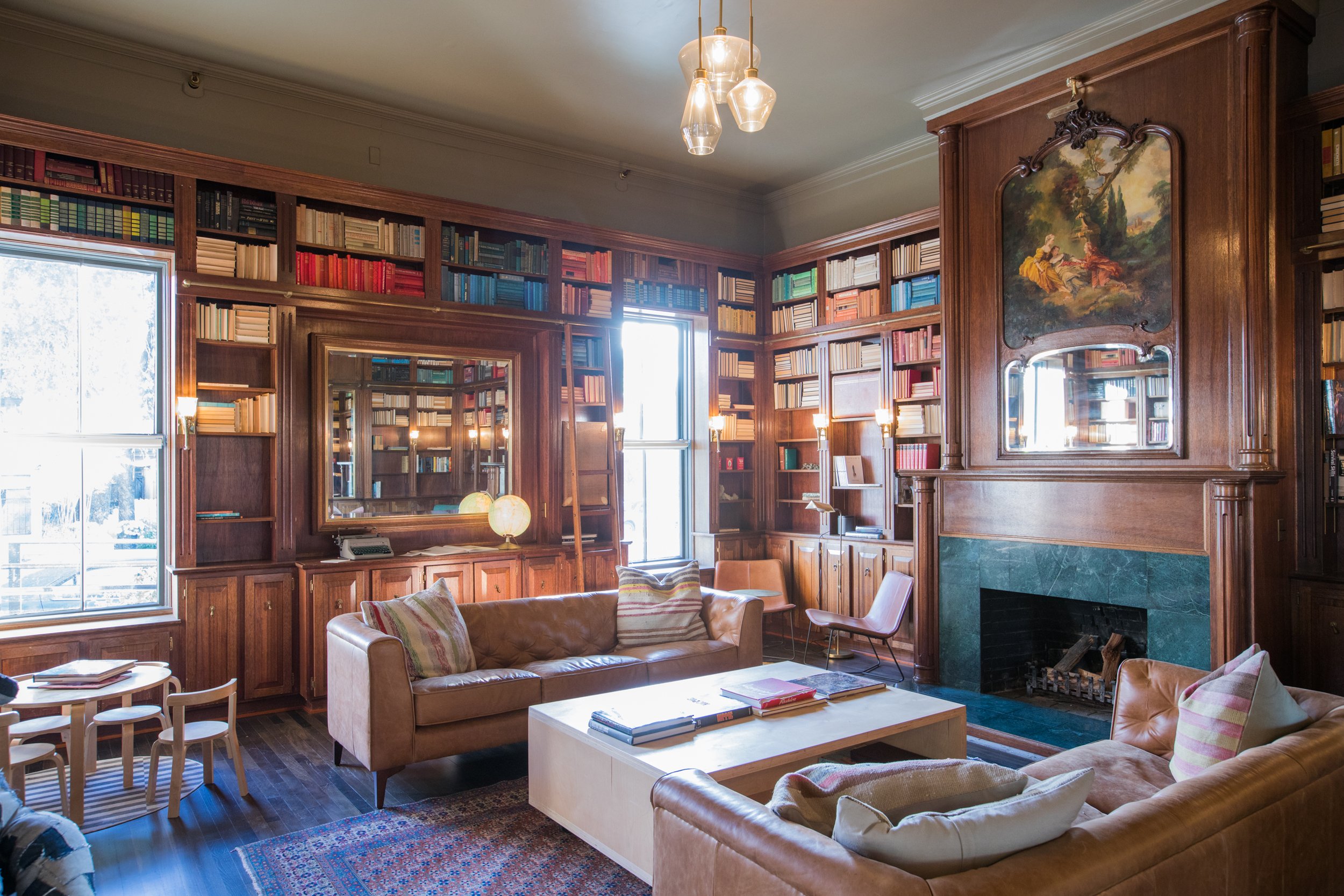 A cozy living room with wooden bookshelves filled with colorful books, a stone fireplace, a tan leather sofa with pillows, chairs, and modern lighting fixtures.