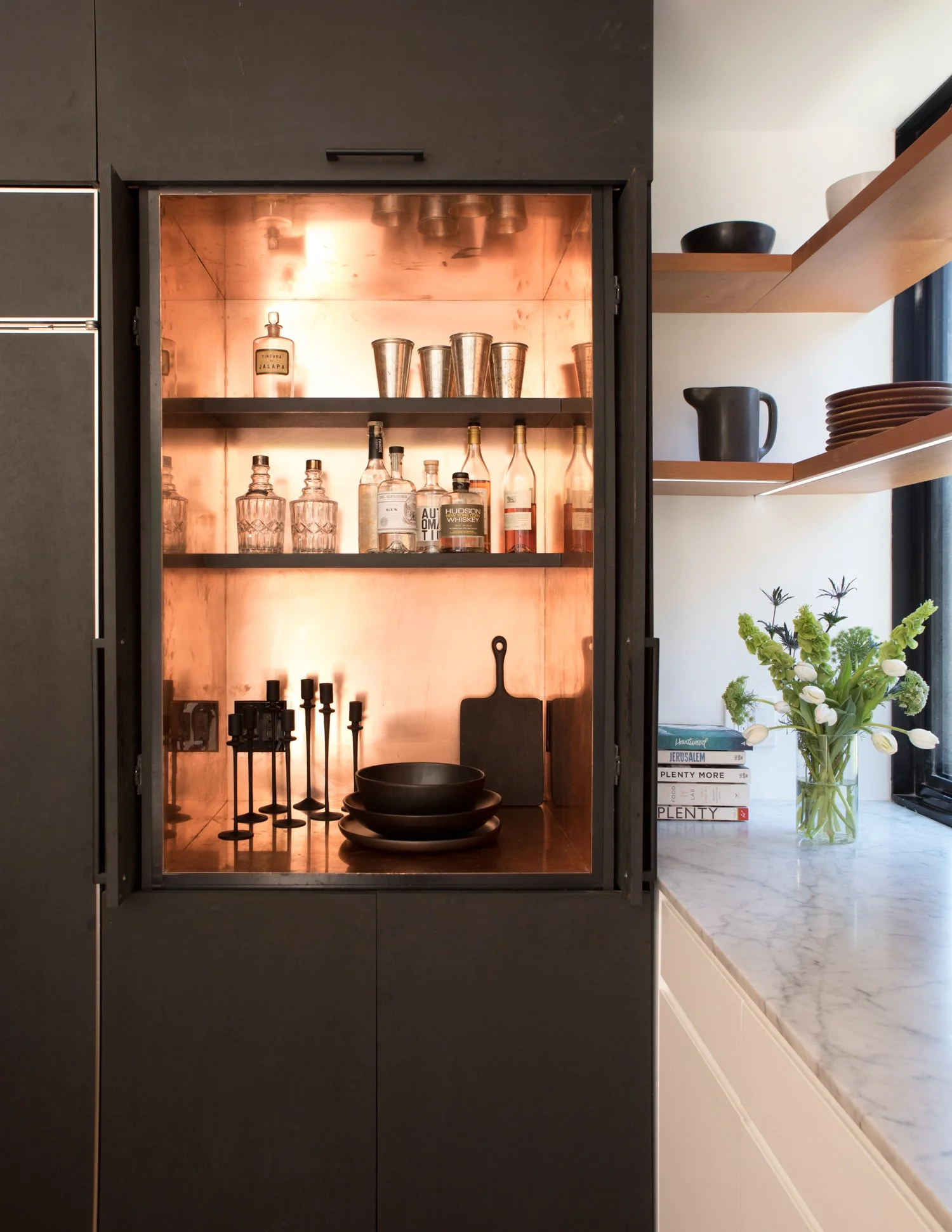 A built-in kitchen cabinet with black doors and a copper-toned interior, containing liquor bottles, glasses, and black dishware, with open shelves holding black bowls, plates, a pitcher, and books, alongside a flower arrangement on a marble counterto