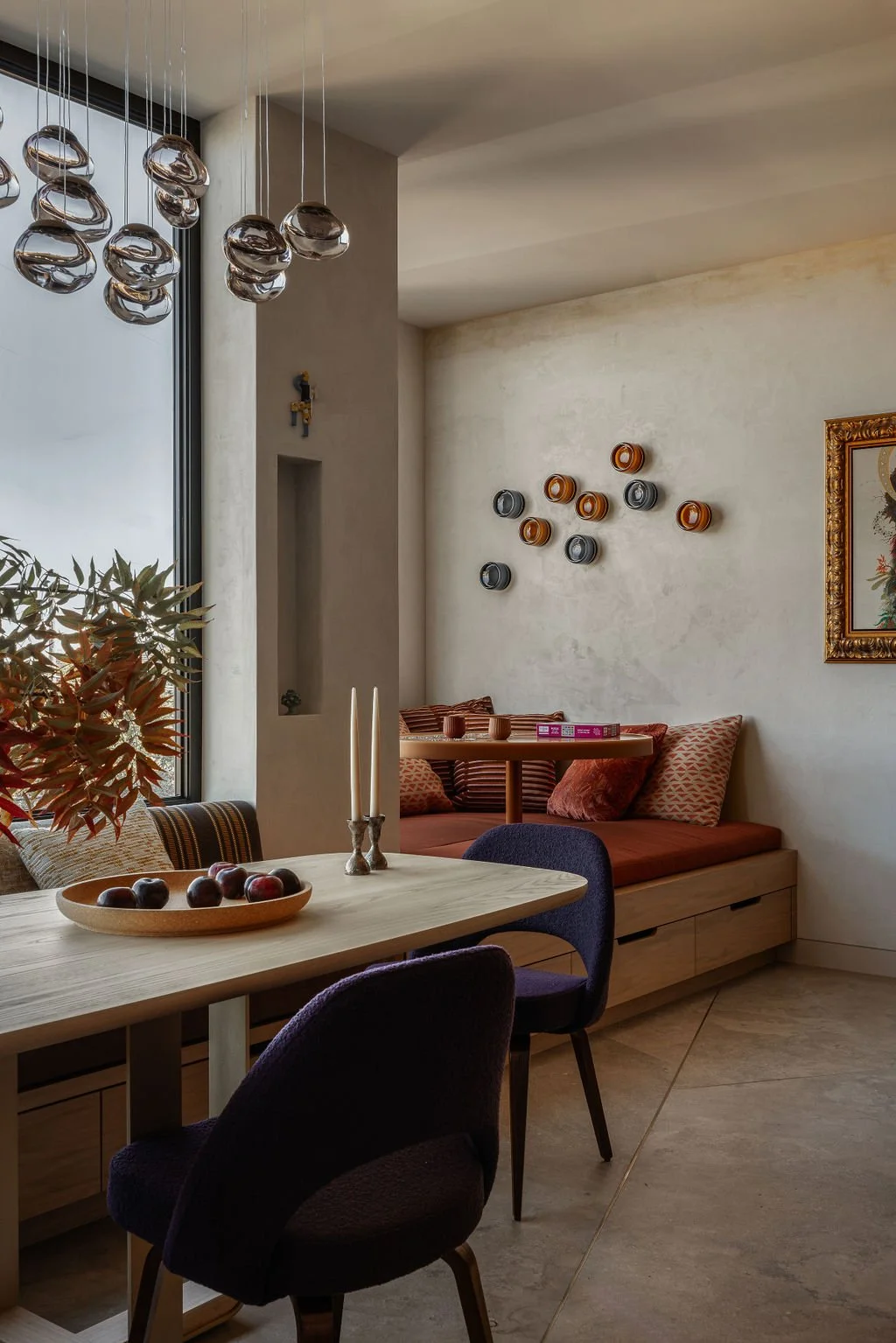 Modern dining room with a light wooden table, dark chairs, a window with potted plant, a built-in bench with red cushions and pillows, decorative wall art, hanging pendant lights, and a framed picture on a textured wall.