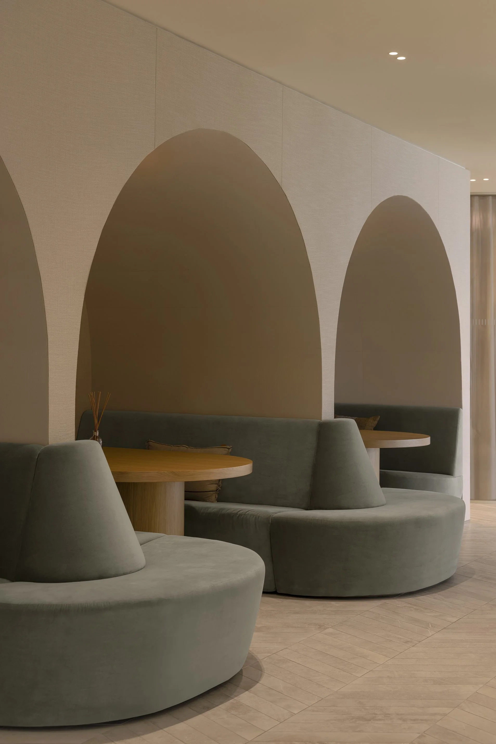 Modern interior with curved green sofas and round wooden tables, separated by a wall with arched openings, in a softly lit space.