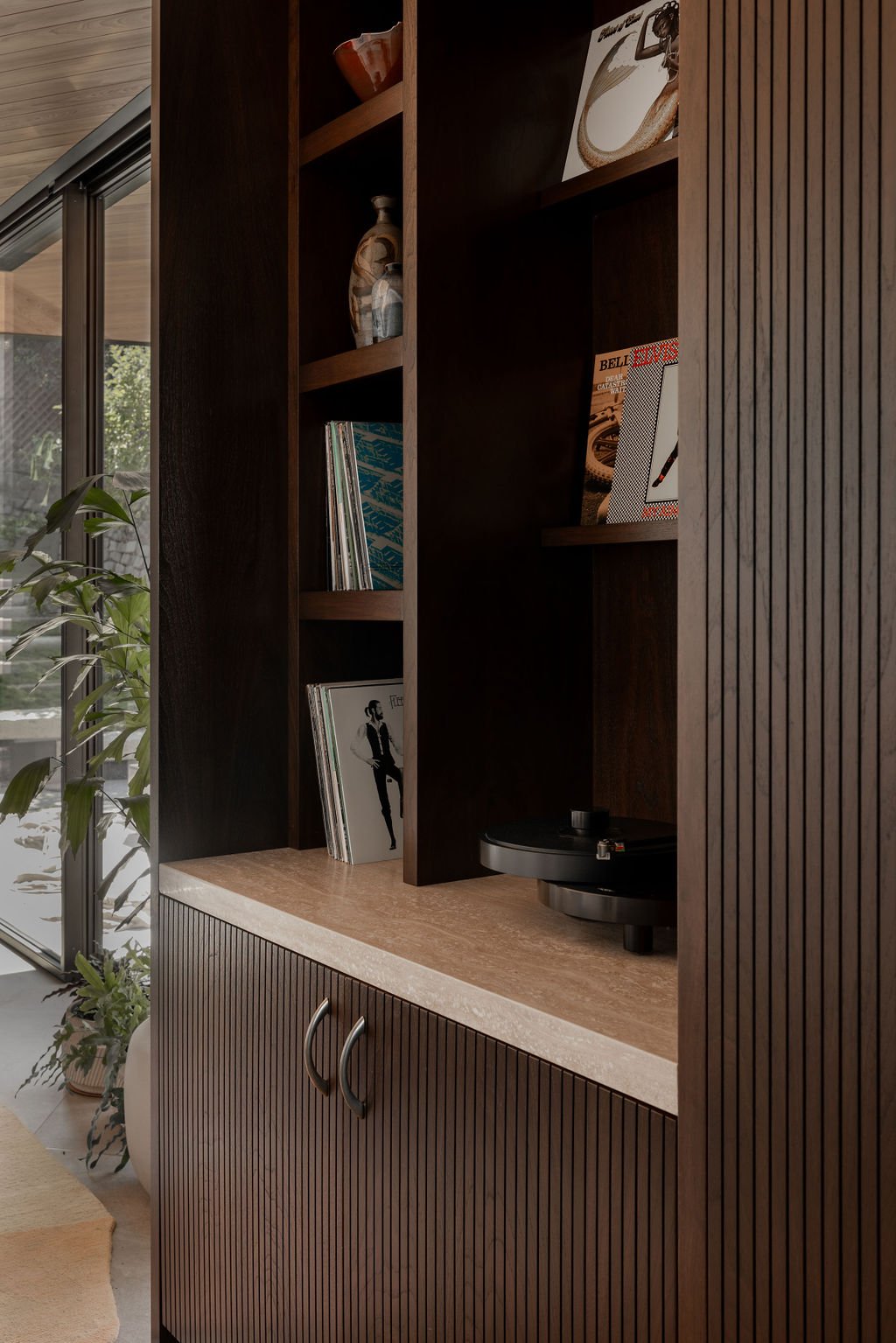A wooden bookshelf containing vinyl records and magazines, with a turntable on the bottom shelf, next to a large window with plants outside.