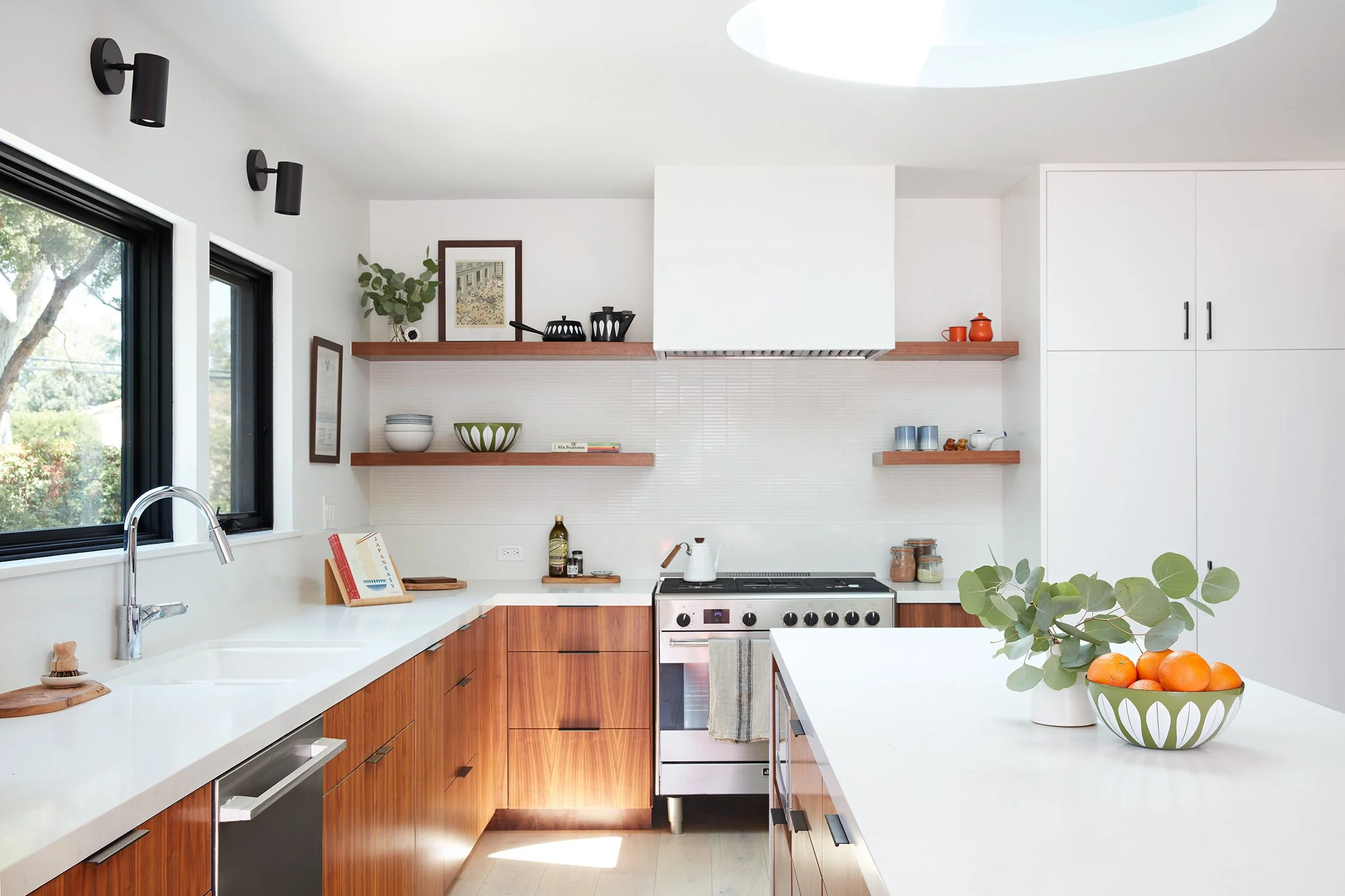 Modern kitchen with white countertops, wooden lower cabinets, and open shelving with dishes, a plant, and decorative items. Large window, black wall lights, and a stove with a tea kettle. Bright, minimalist design with natural light.