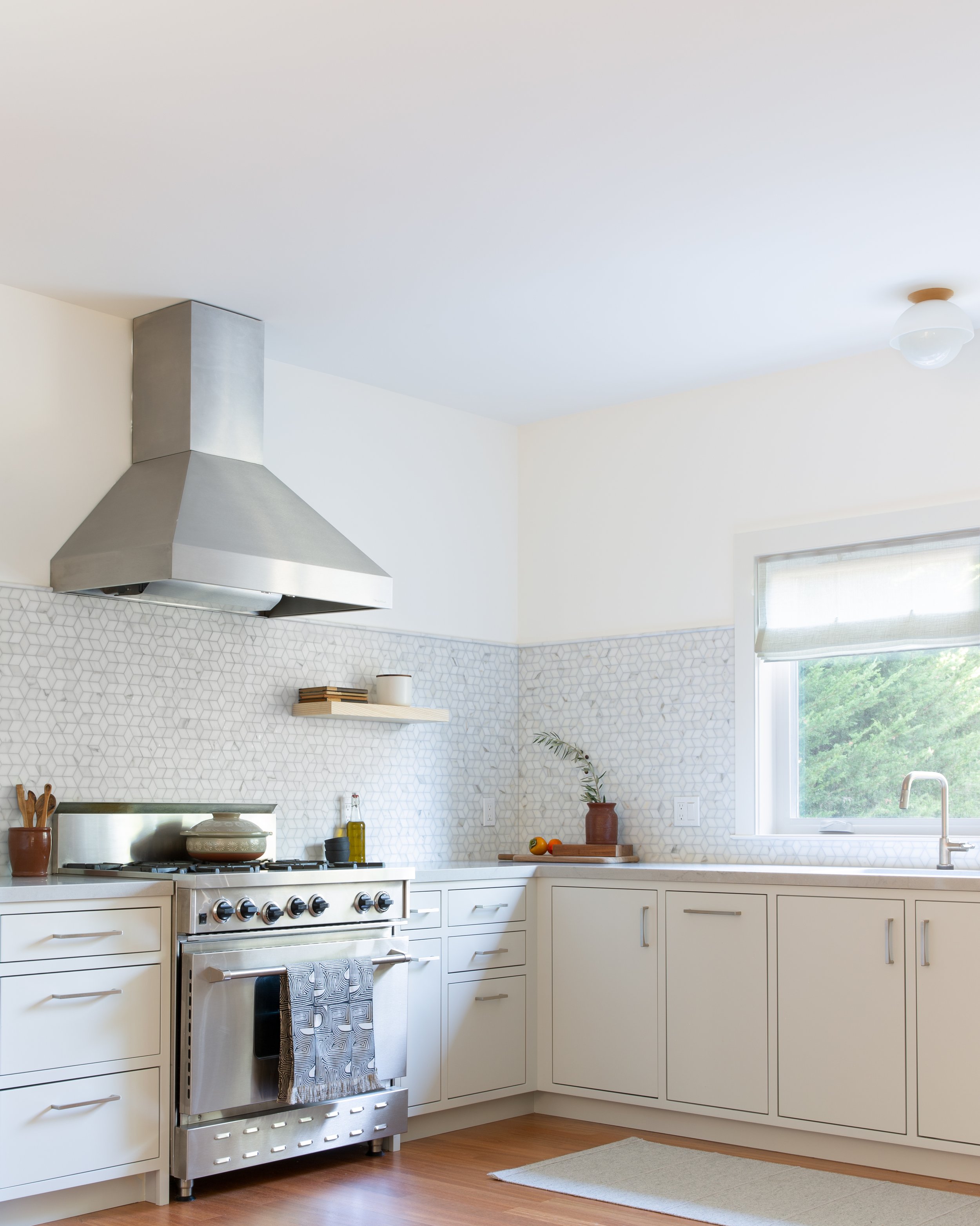 Modern kitchen with white cabinets, stainless steel stove and range hood, white geometric patterned backsplash, window with blinds, small potted plant, and a countertop with fruit and oil bottles.