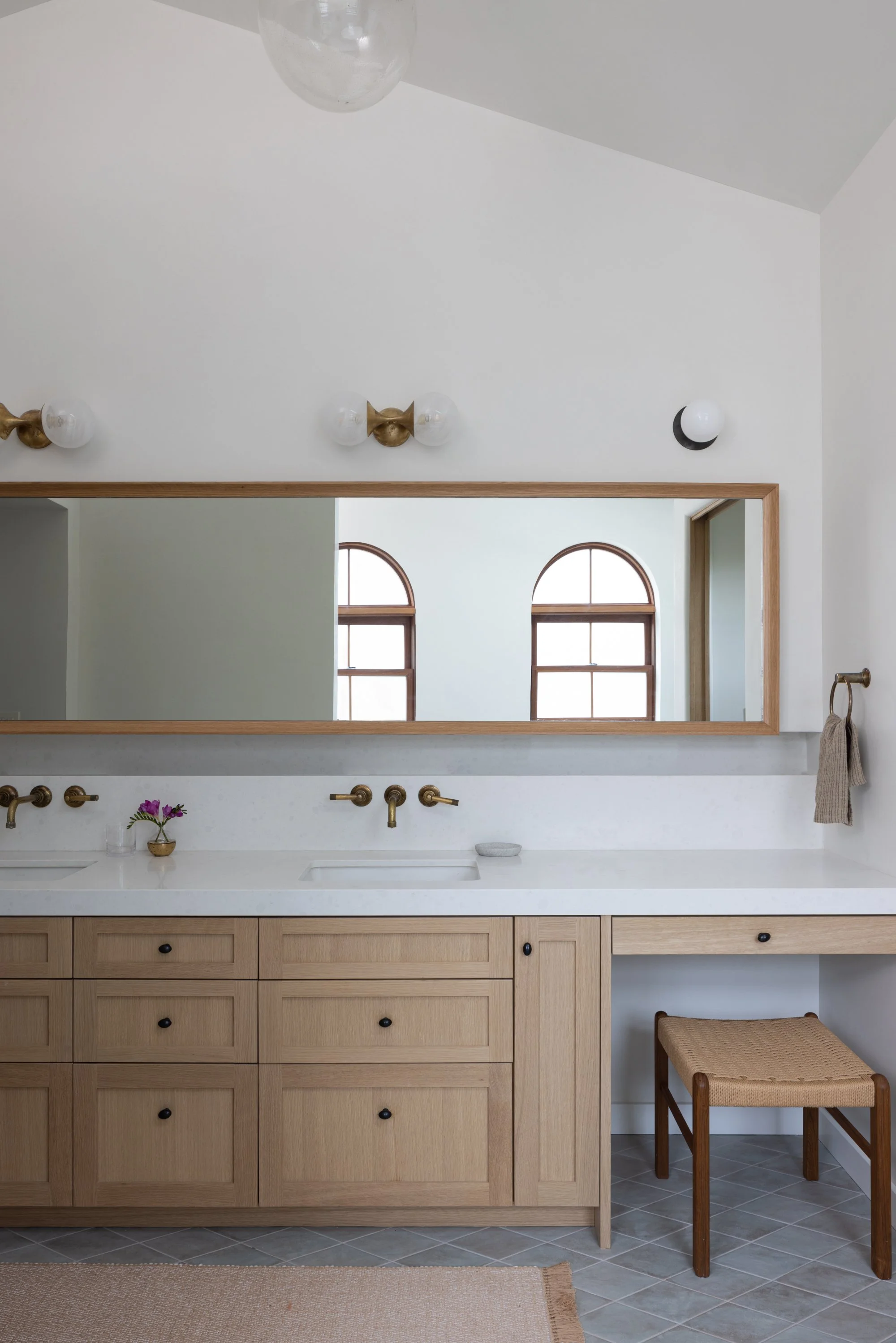 A bathroom vanity with a white countertop, light wood cabinets, a large rectangular mirror, brass faucets, and a small potted flower. There is a woven stool and a towel on a hook next to it. The background has two arched windows, and the wall feature