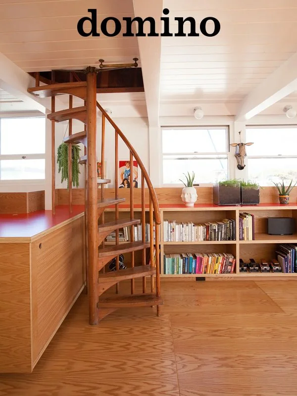Interior view of a room with wood flooring, a spiral wooden staircase, a built-in bookshelf filled with books, plants, and decorative items under two large windows.