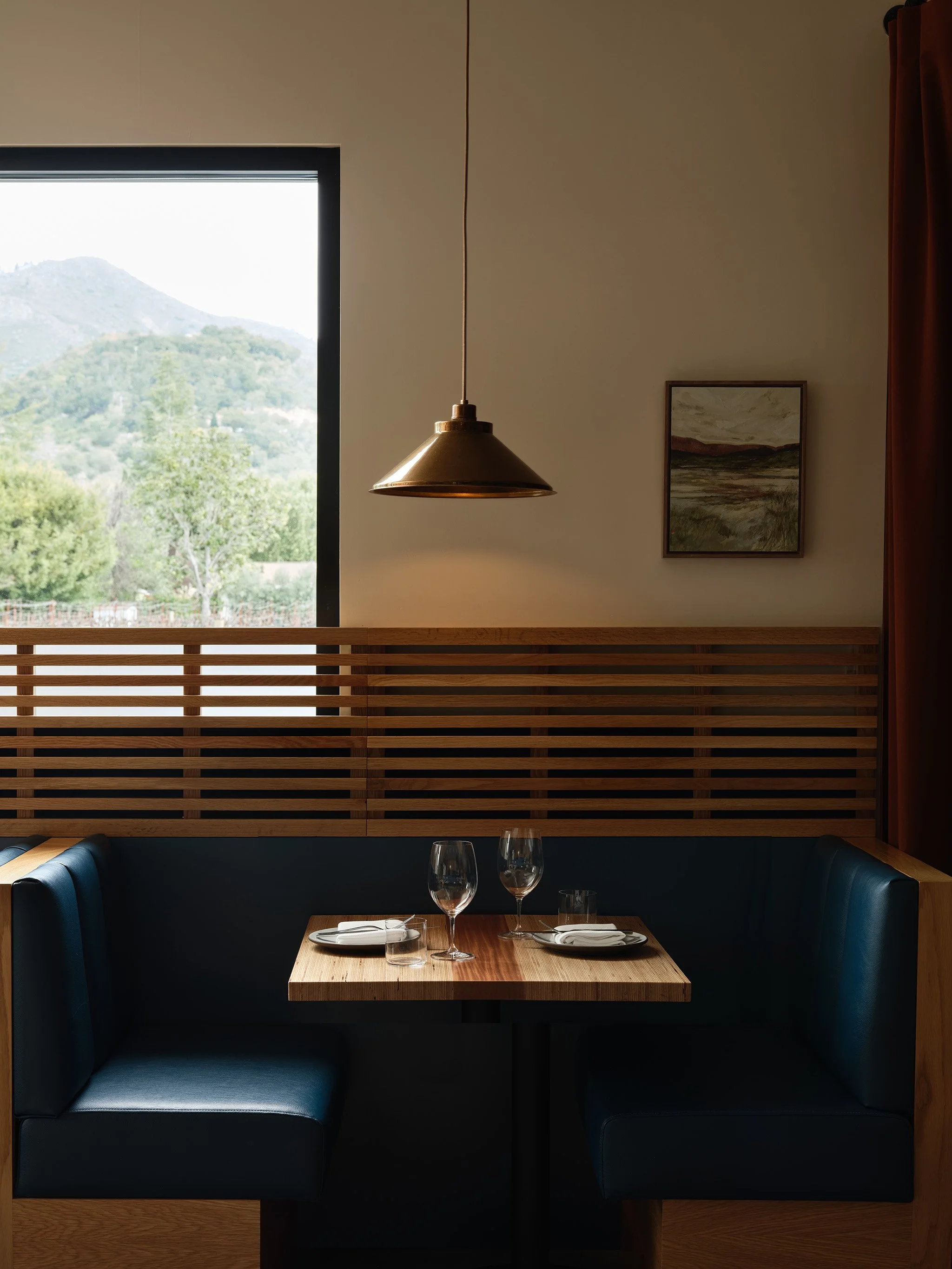 A restaurant booth with dark blue cushioned seating, a wooden table set with plates, napkins, silverware, and three wine glasses, framed landscape art on the wall, a window with a view of green trees and distant mountains, and a hanging brass pendant