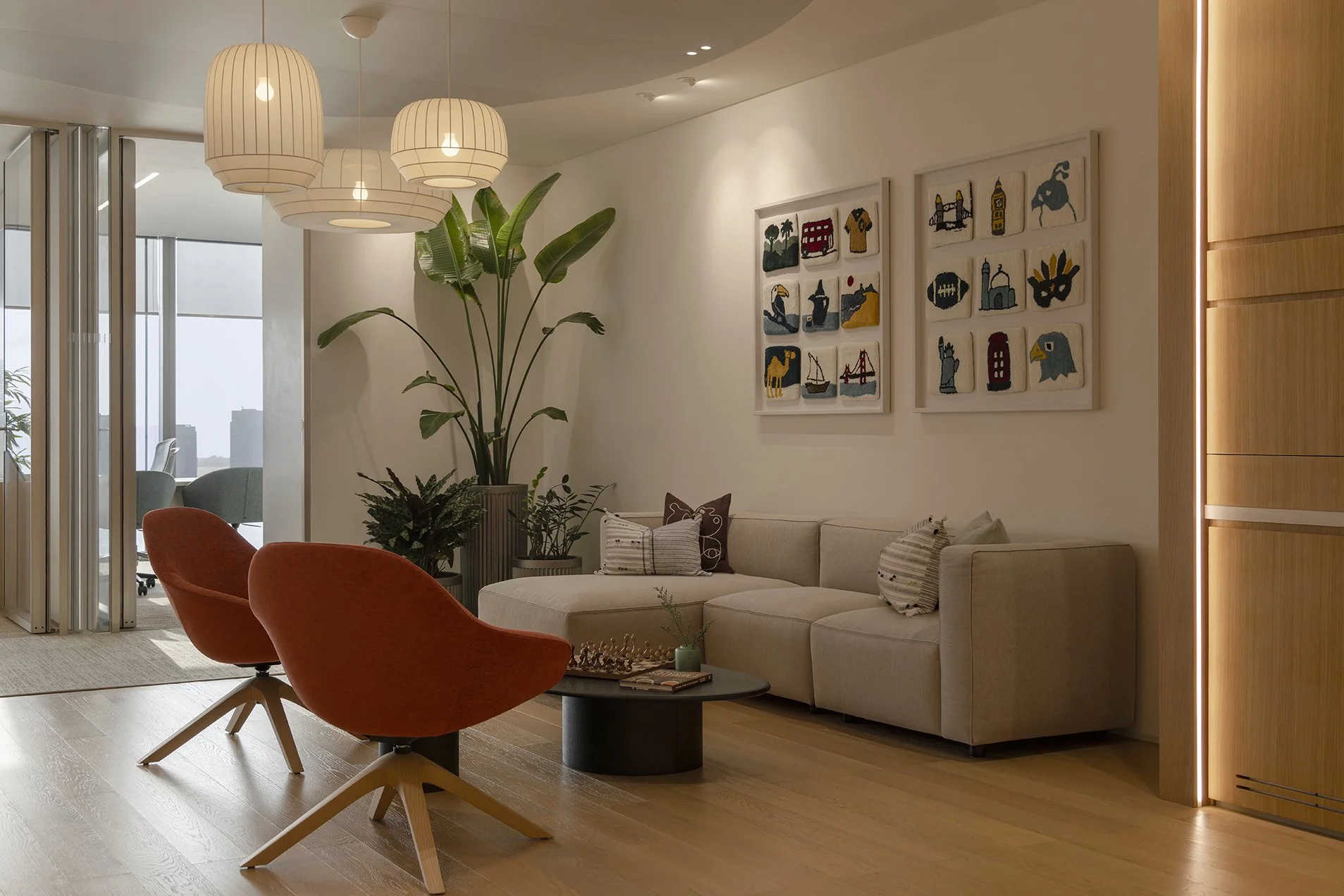 Modern living room with beige sofa, orange and patterned chairs, large indoor plants, wall art, wooden flooring, and pendant lights.