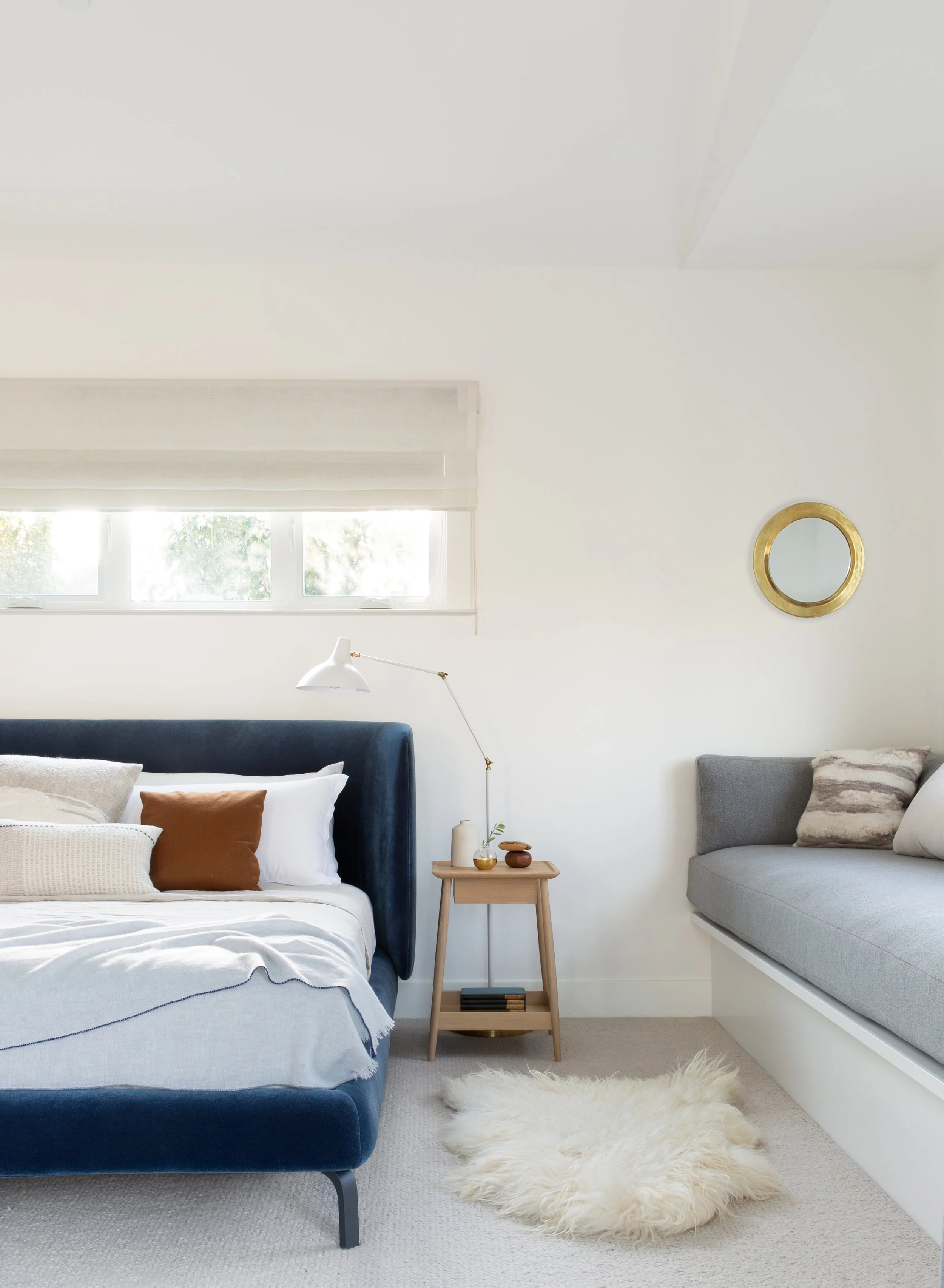 A bedroom with a white bed, dark blue headboard, and white bedding with pillows. Next to the bed is a small wooden bedside table with decorative vases. There is a window with a white shade, a white wall with a round gold-framed mirror, a gray window 