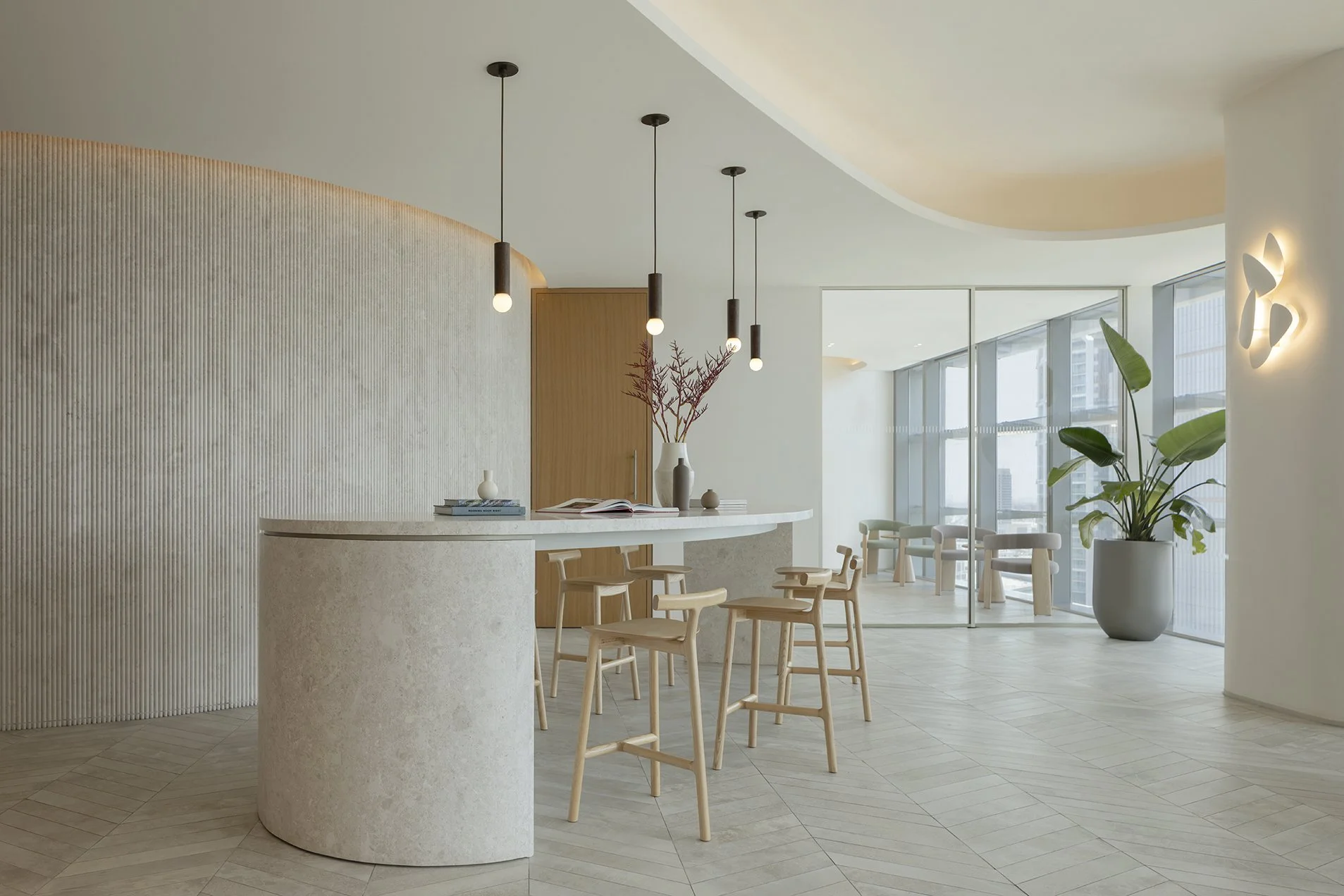 Modern interior with a curved reception desk, light wood chairs, pendant lighting, large green plant, and floor-to-ceiling windows in a bright space.