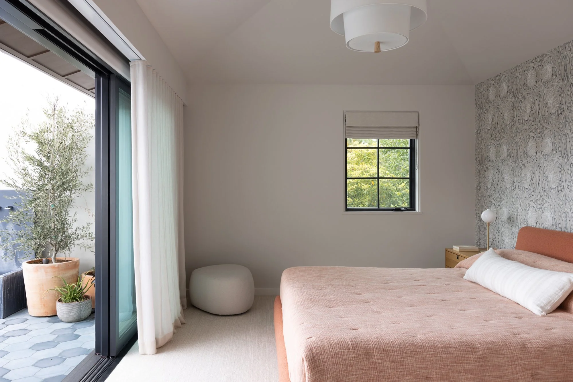 A cozy bedroom with a large bed, pink bedding, a nightstand with a lamp, a small window showing green trees, and an open sliding door leading to an outdoor area with potted plants.