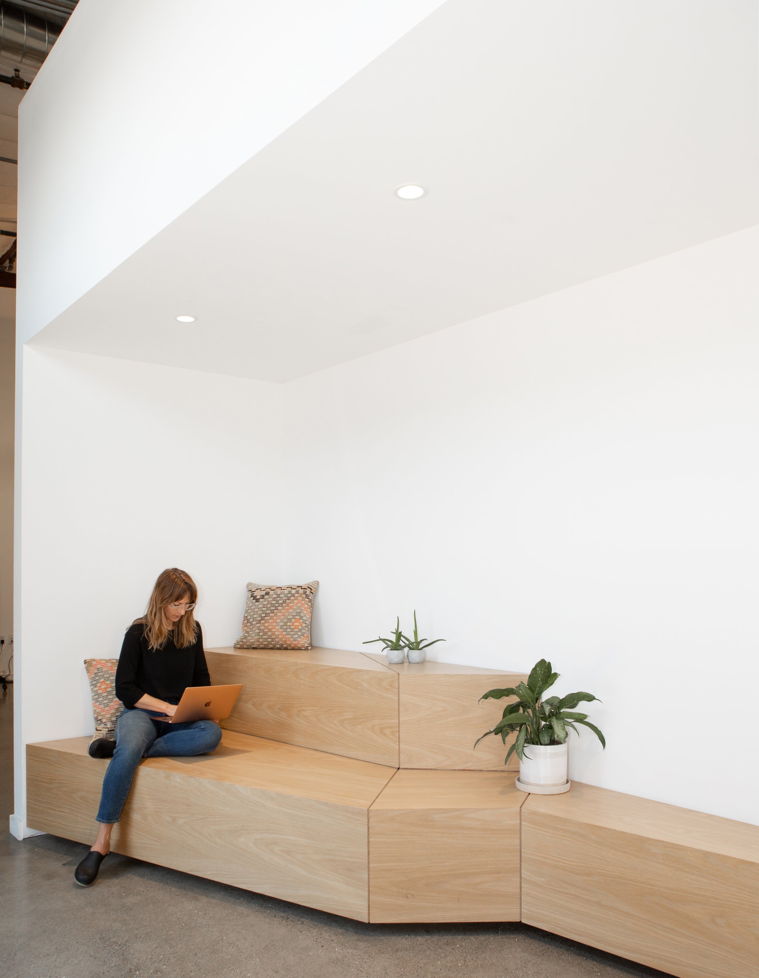 Woman sitting on wooden steps with laptop, leaning against a white wall, with cushions and potted plants nearby.